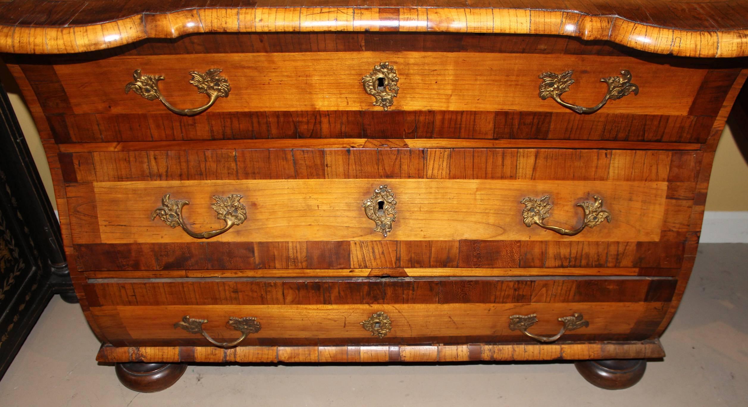 A German walnut commode, featuring a conforming crossbanded top with contrasting fruitwood inlay and molded edges, surmounting a bombe case with three shaped drawers, each with cross banded fronts, original hardware, and contrasting marquetry side