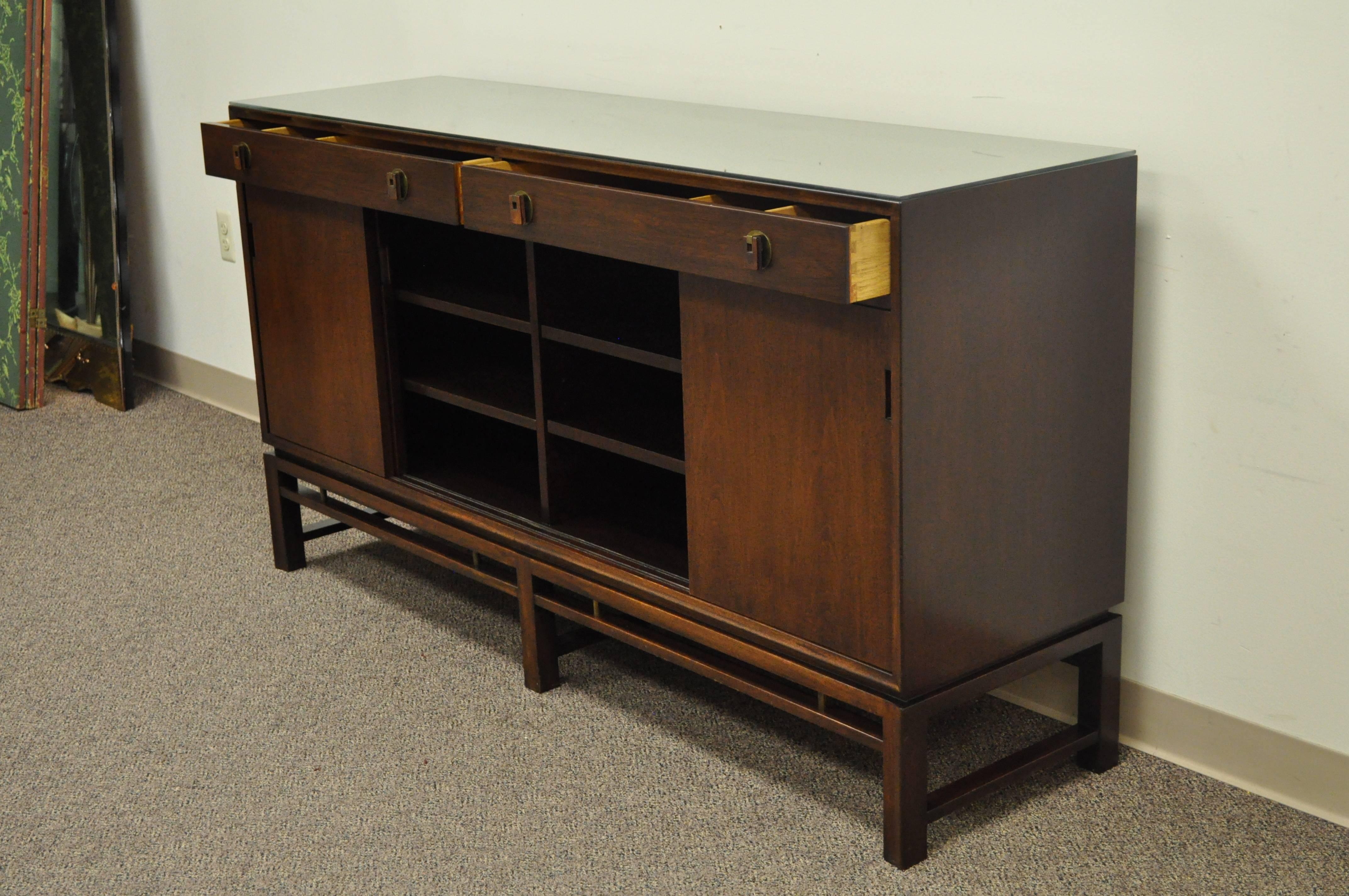 Edward Wormley for Dunbar mahogany sideboard. Item features clean Mid-Century Modern lines, brass accents at the base, two dovetail constructed upper drawers. four sliding cabinet doors, brass and mahogany pulls, and a custom glass top.