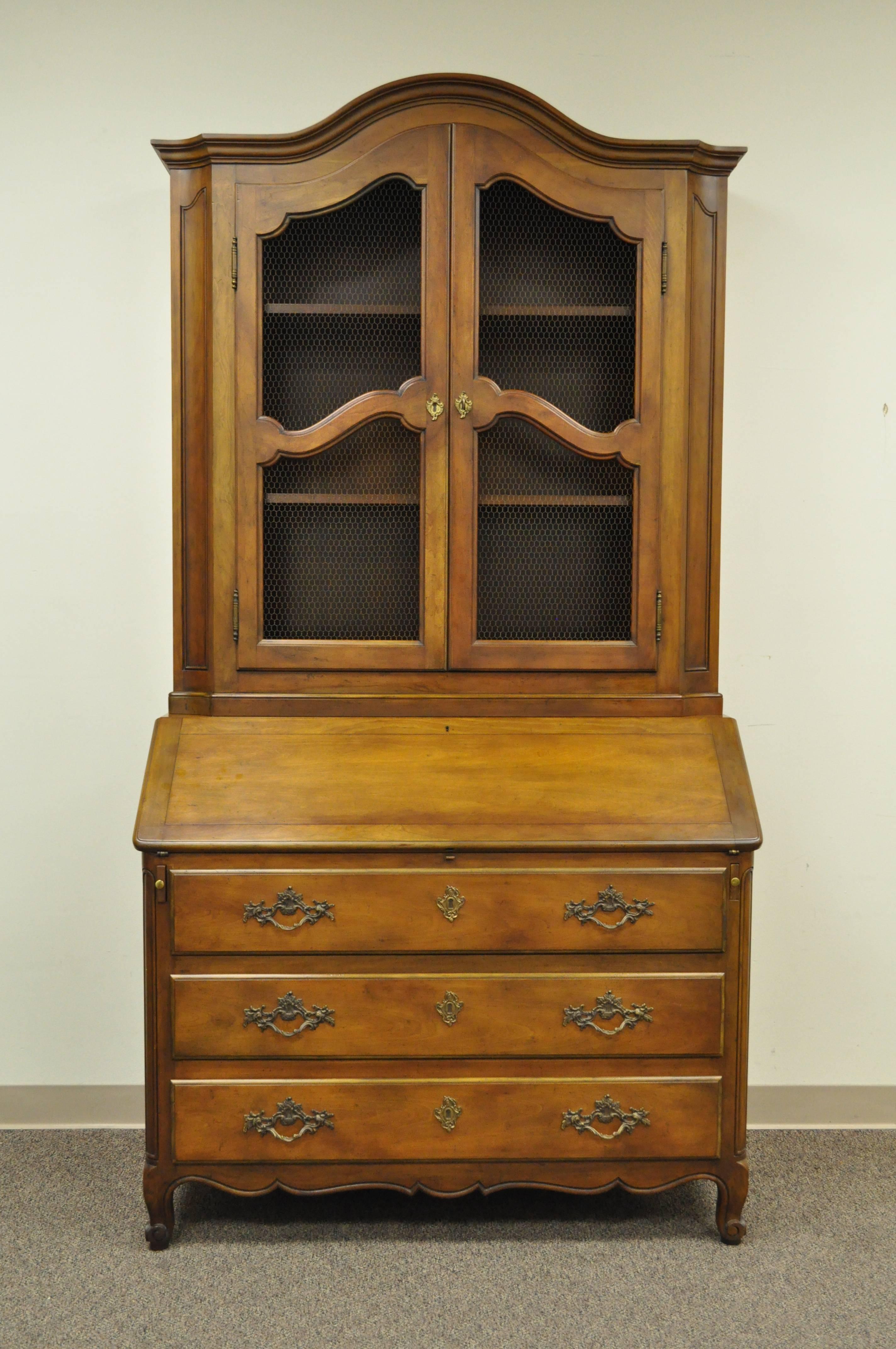 Remarkable quality two-piece walnut secretary desk by Baker Furniture Co. in the Country French Taste. Item features a bonnet top bookcase upper with adjustable interior shelving, metal lattice door fronts, drop front writing desk with leather top