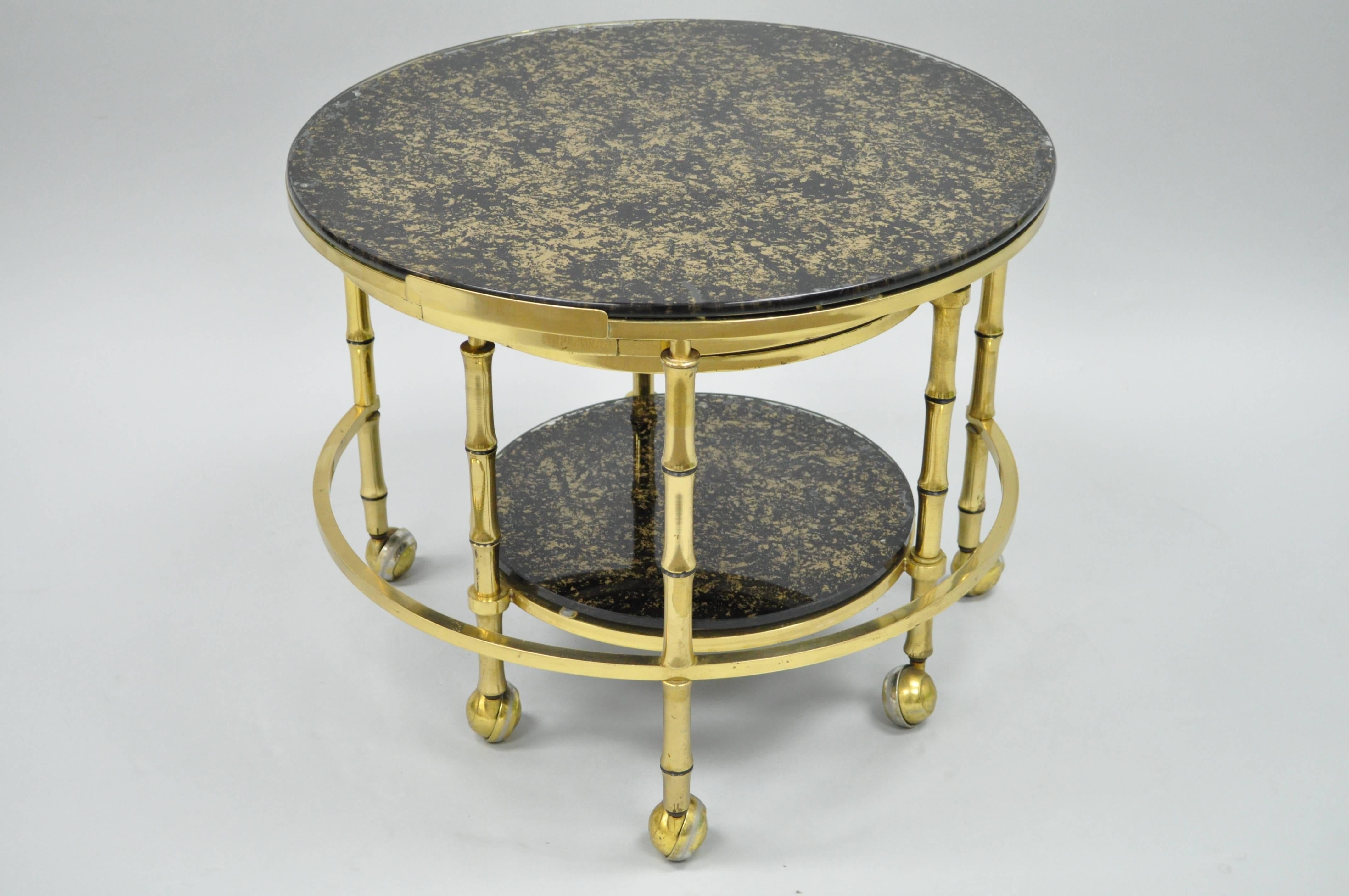 Vintage Hollywood Regency faux bamboo solid brass and glass nesting tables. This unique set features three round brass faux bamboo folding/expanding frames on rolling casters and gold flecks decorated black glass tops. Great for use as a coffee