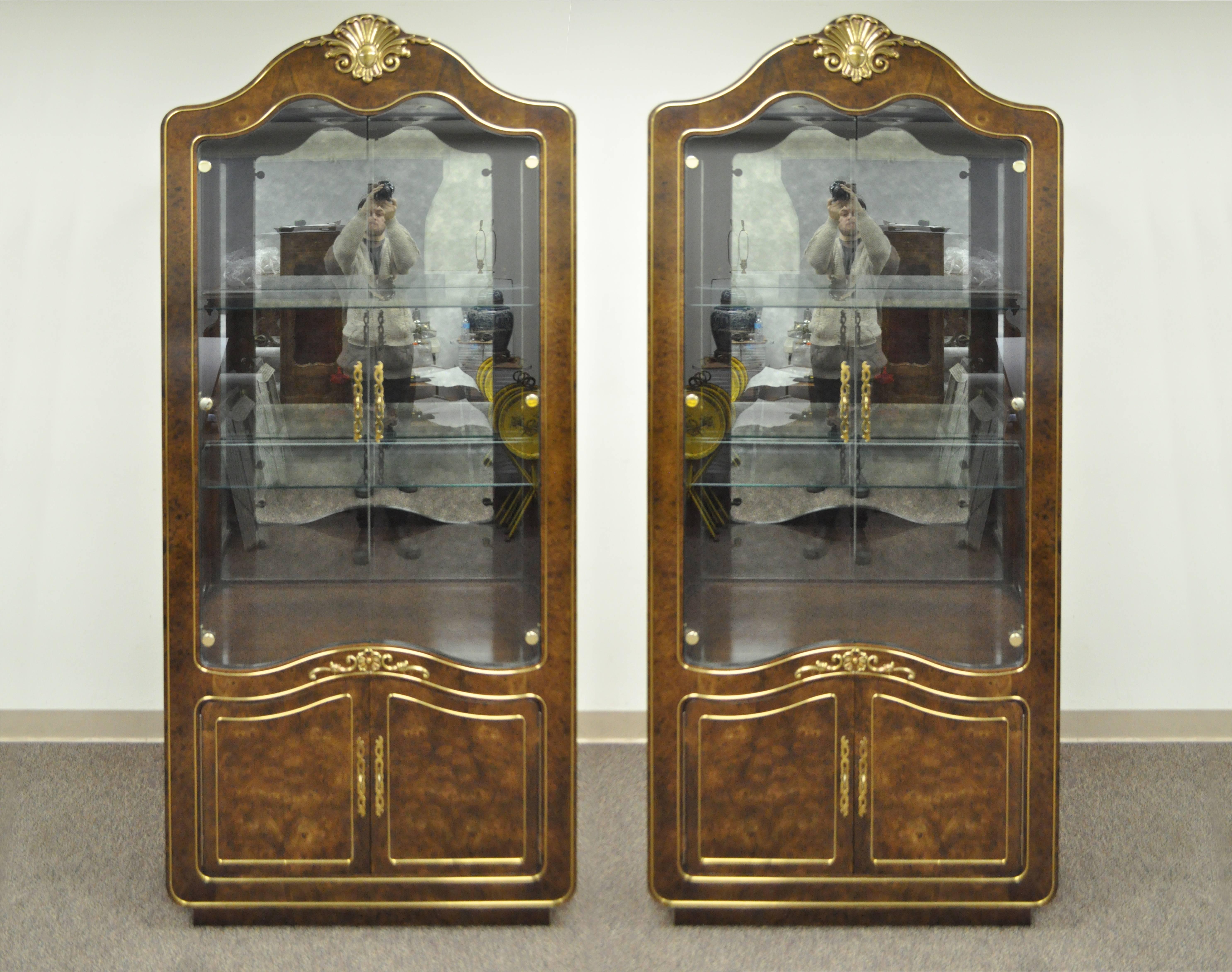 Remarkable pair of Amboyna burl wood French Hollywood Regency style curio display cabinets by Mastercraft. Each etagere cabinet / bookcase features a lighted interior with dimmer, brass inlaid trim, brass central shell form crest, bonnet top, two