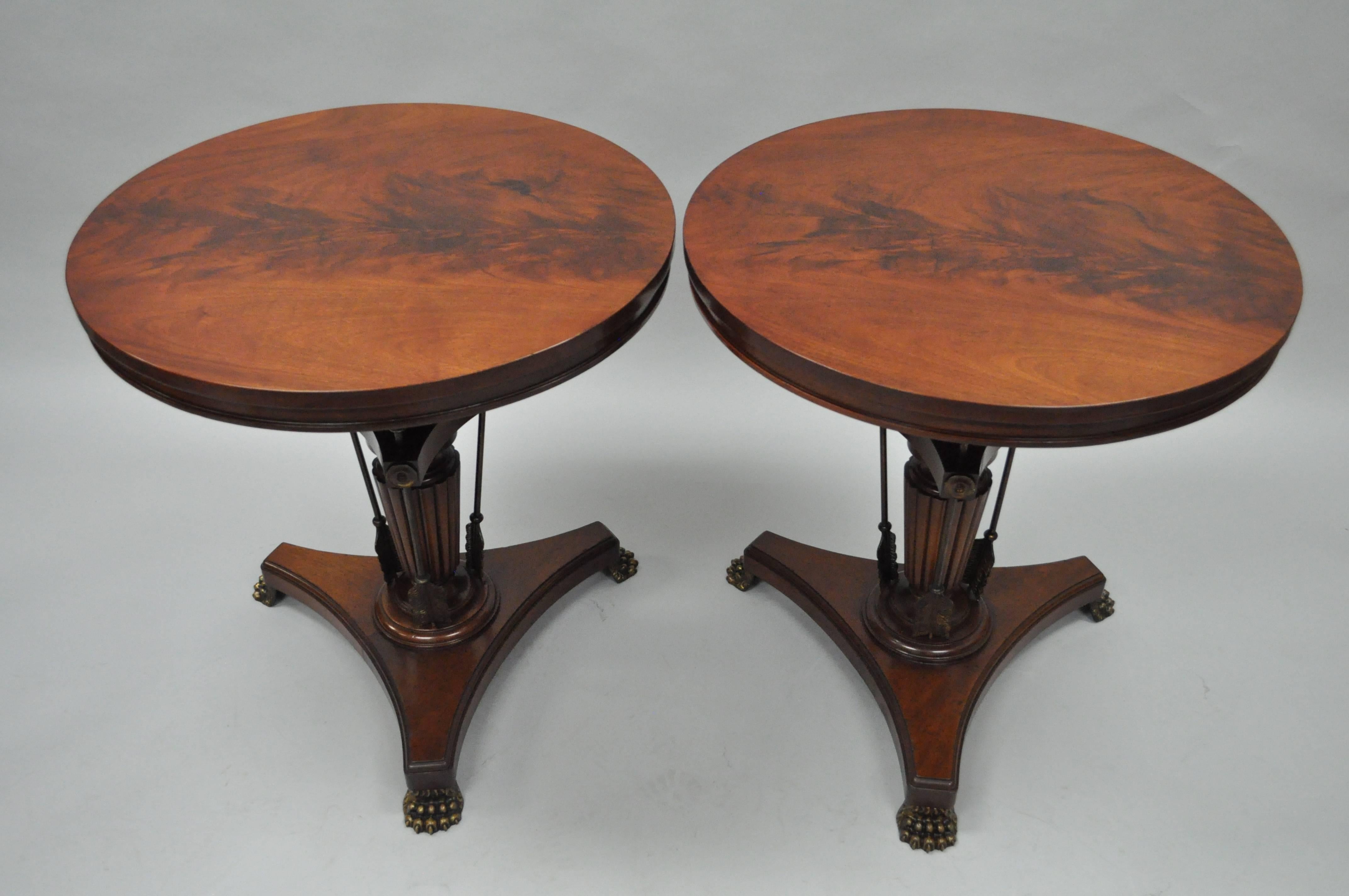 Quality pair of crotch mahogany and brass Neoclassical style arrow base round side or accent tables. Item features figured crotch mahogany tops over solid mahogany reed carved pedestal bases with triple brass arrows and cast brass paw feet. Great