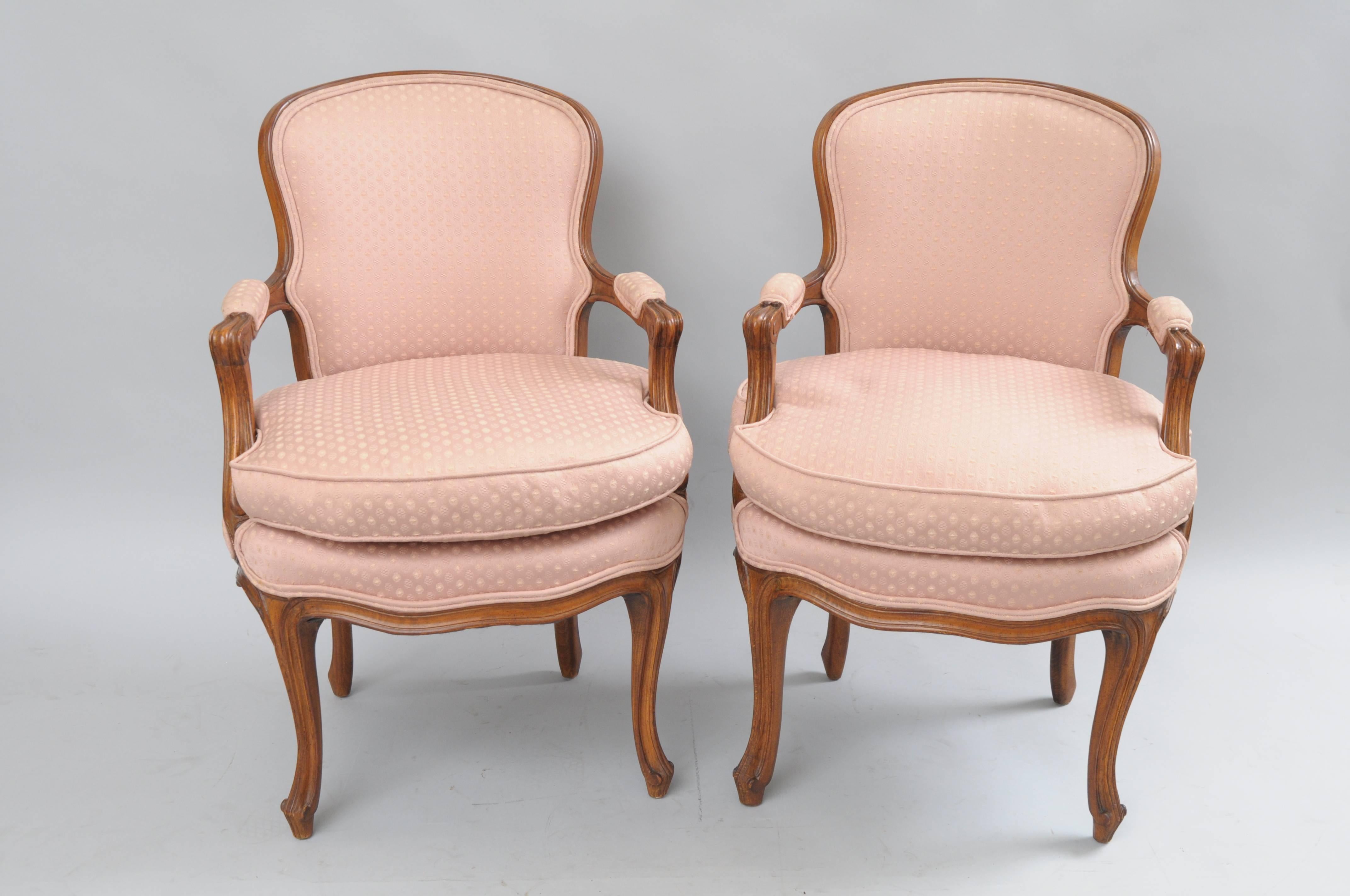 Pair of vintage Country French / Louis XV Style carved walnut living room / bedroom boudoir arm chairs. The pair features uniquely shaped solid carved wood frames, rounded upholstered backs and armrests, shapely cabriole legs, shaped lower rails,