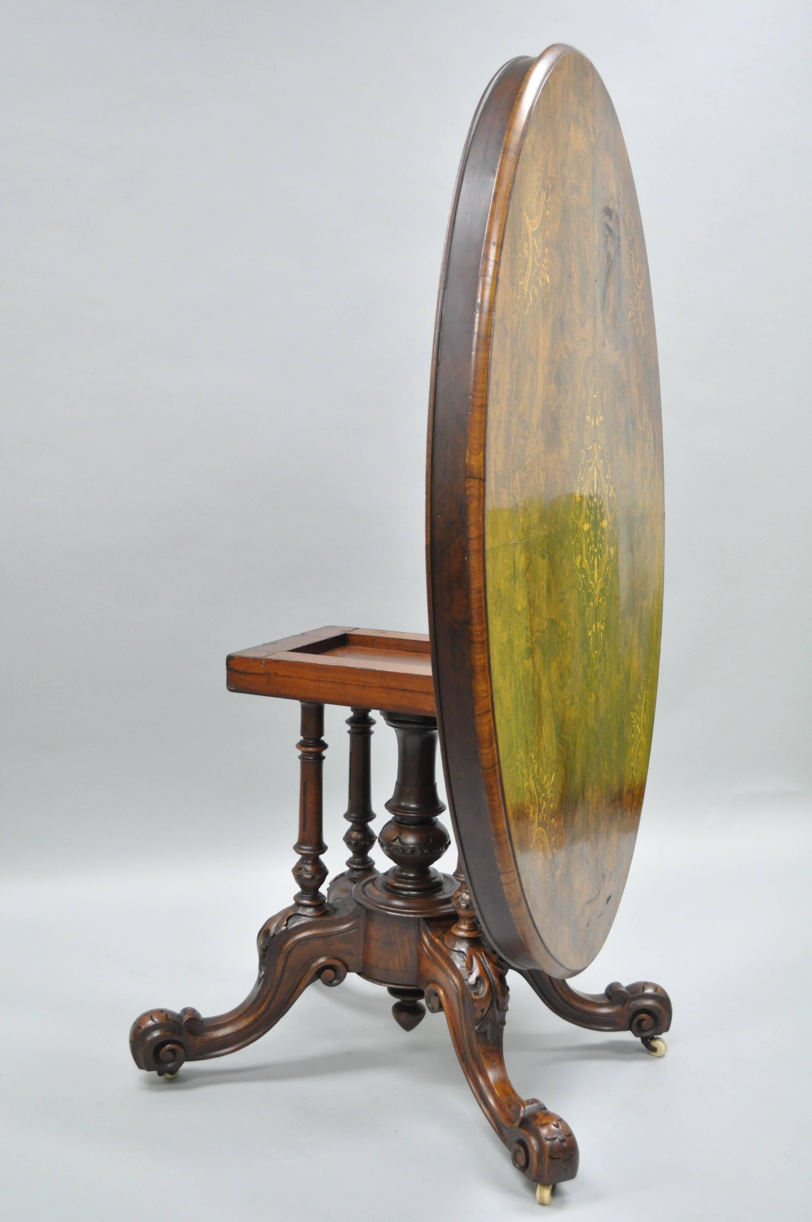 Victorian walnut tilt-top center table, circa late 19th century. The oval burl wood top, inlaid with floral marquetry, rising on four turned supports with a carved central shaft, foliate carved outswept curved legs on scroll feet and casters. 62