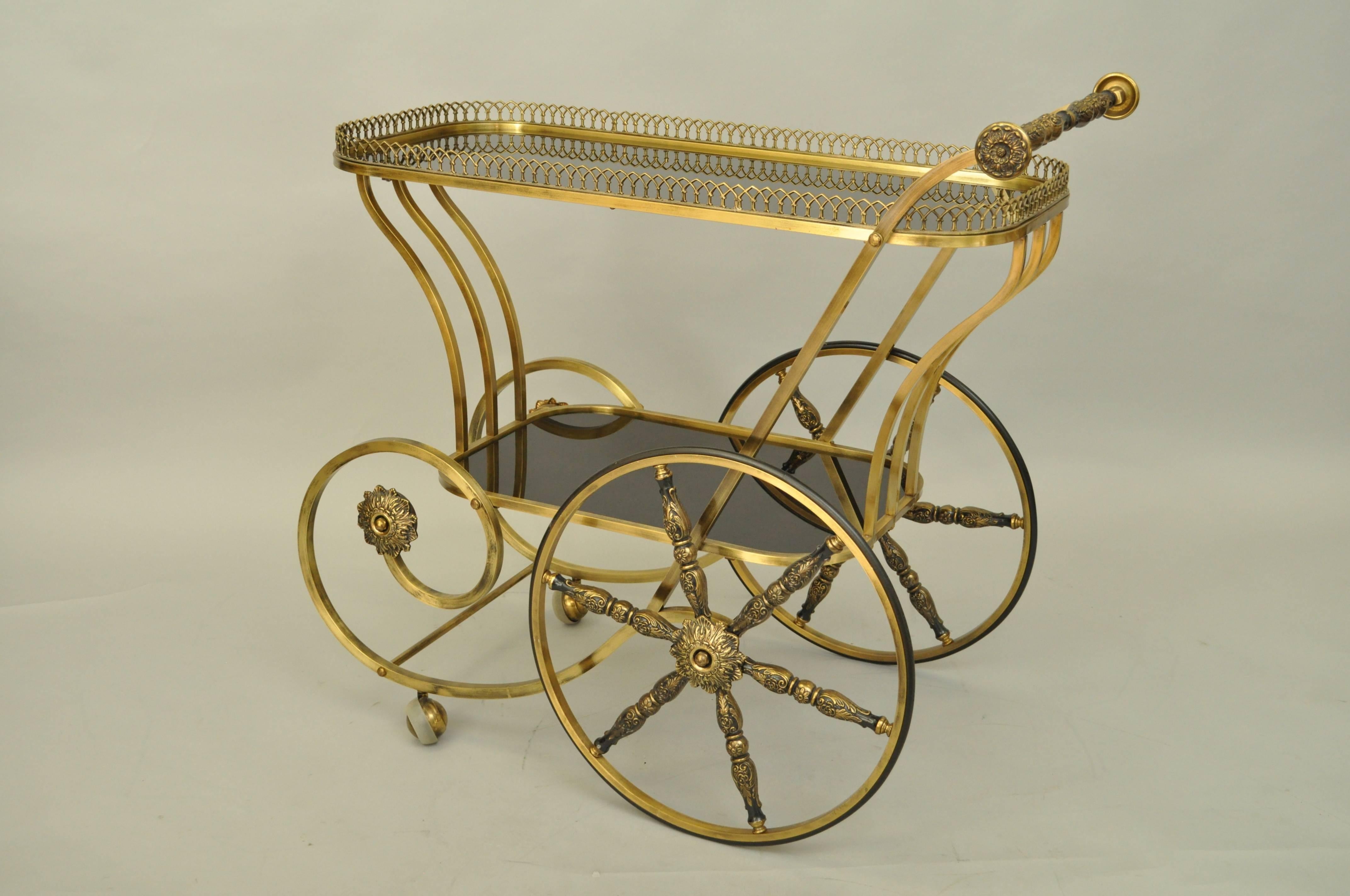 Very unique vintage two-tiered bar cart featuring a scrolling brass frame, ornate wheel spokes and handle, pierced brass gallery and two-tiered of deep gold finished glass.
  