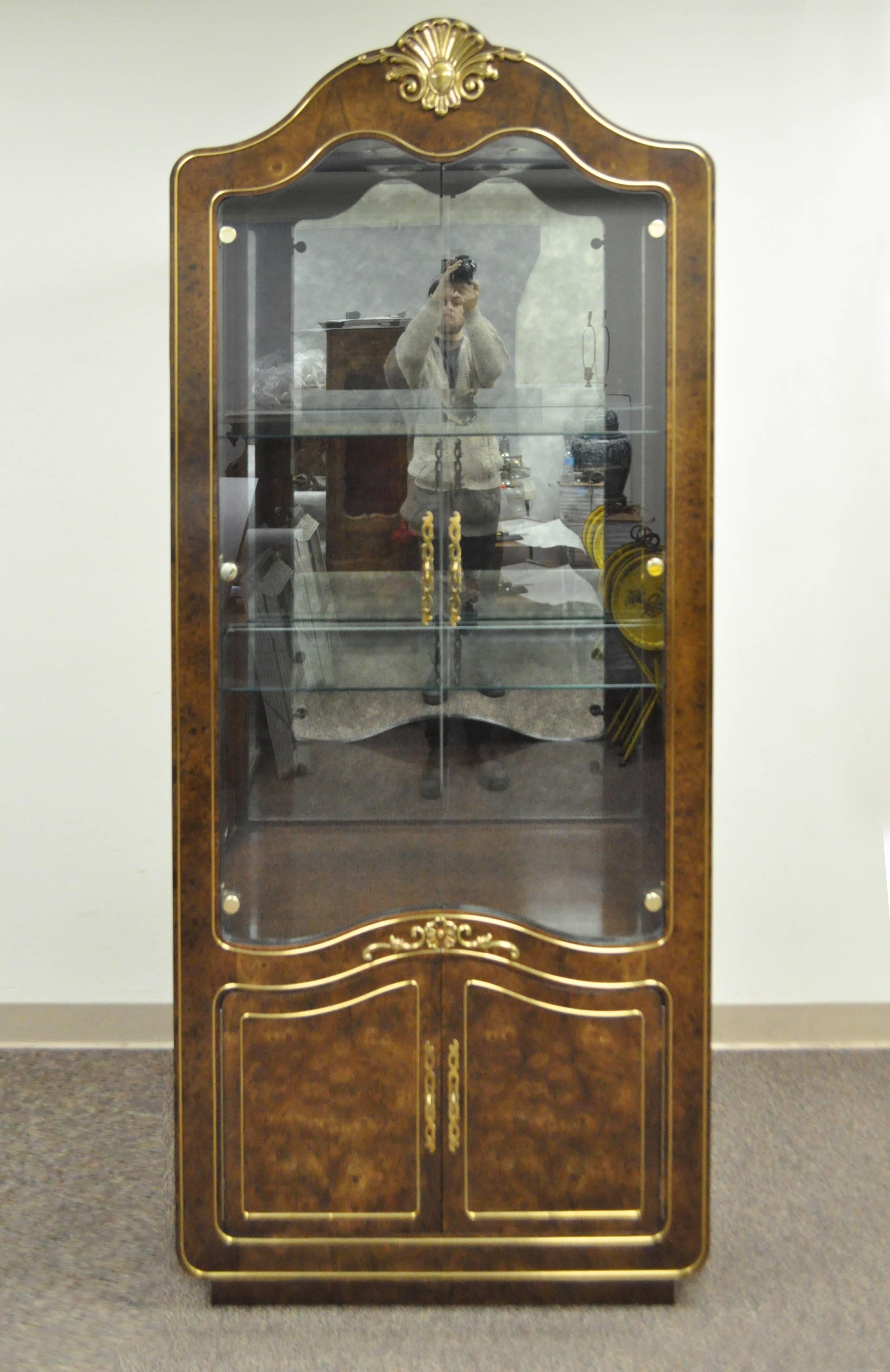 Remarkable Amboyna burl wood French Hollywood Regency style curio display cabinet by Mastercraft. Etagere cabinet / bookcase features a lighted interior with dimmer, brass inlaid trim, brass central shell form crest, bonnet top, two swing clear