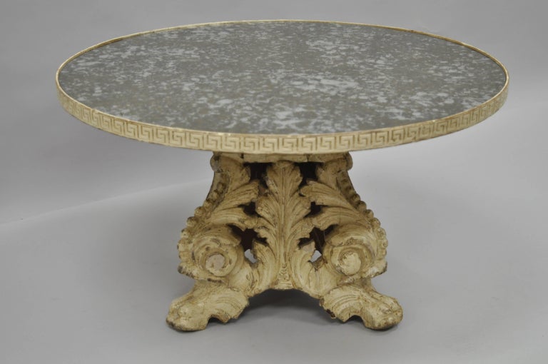 Antique Italian Baroque style round coffee table. Item features an antiqued / églomisé distressed glass top, carved Greek key banding, solid hand-carved wood acanthus and paw foot pedestal base, and a desirable cream distress painted finish. The