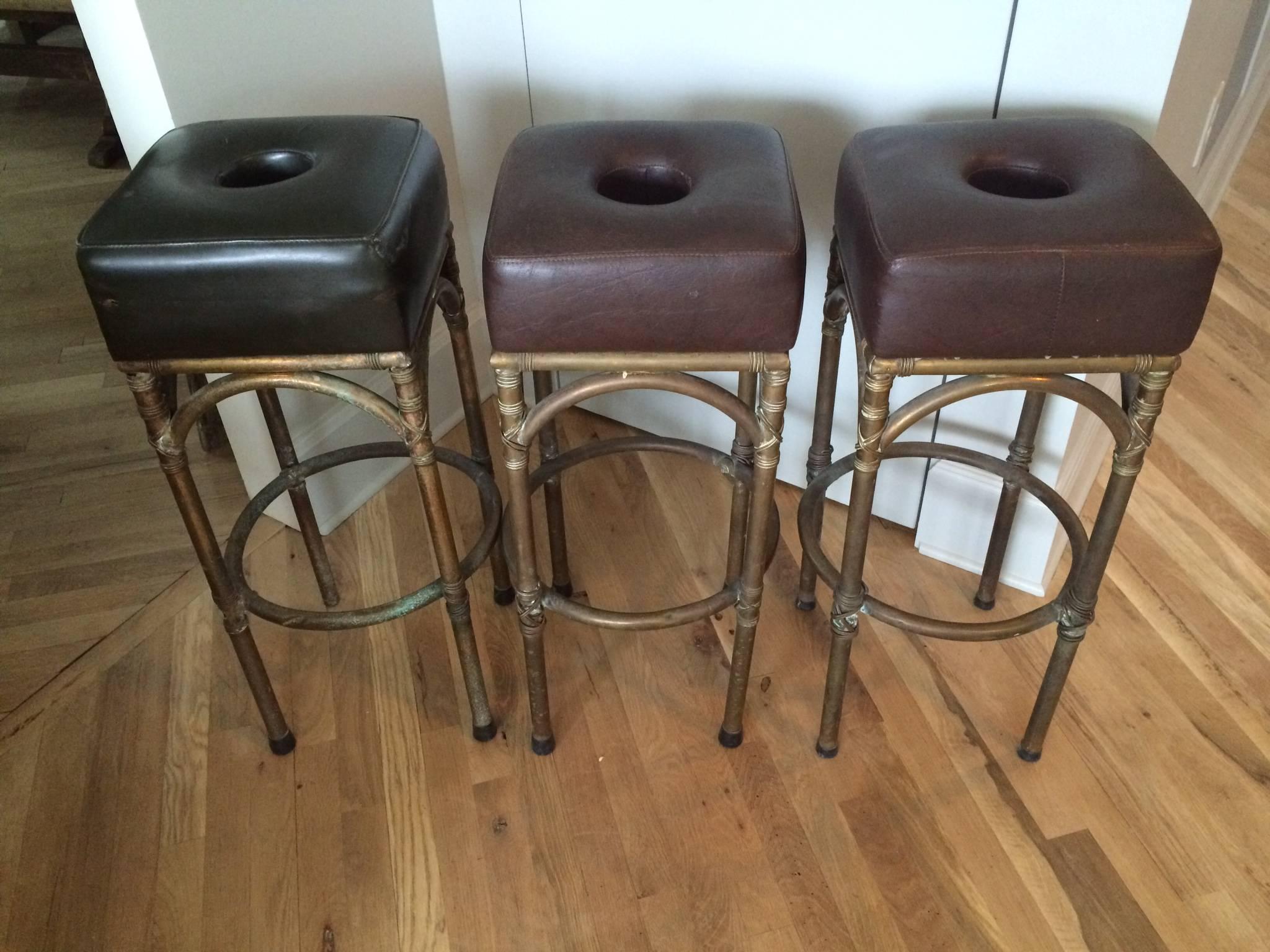 Set of three English leather and brass bar stools from an English pub. Original and quite heavy the cut-out was to reach your hand through and more easily carry the stool. Two are burgundy and one is more green.