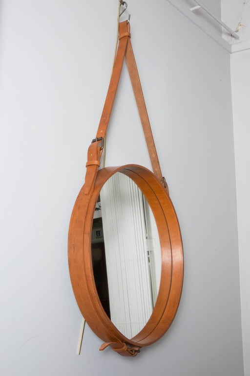 A circular fawn-coloured leather mirror
Held by an adjustable leather strap 
Late 20th century, France
In the style of Jacques Adnet 

Height : 112 cm
Height maximum of the strap : 117 cm 
Diameter : 59 cm
Depth : 8 cm