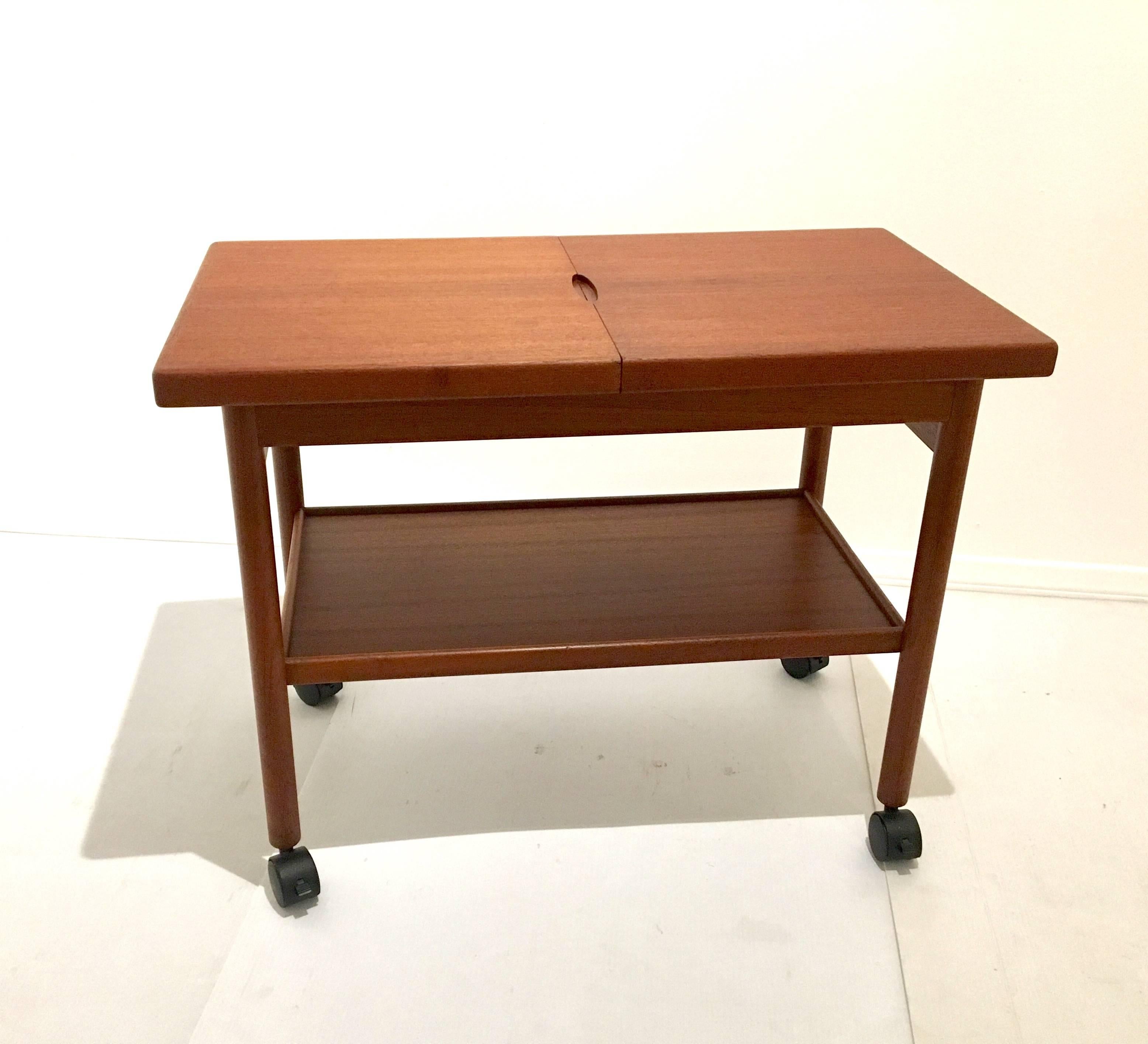 Versatile sliding top serving or bar cart designed by Kurt Ostervig. Top slides open to reveal black melamine top for placing beverages while protecting the teak wood top. A lower shelf for storage the cart has been refinished and each caster has a