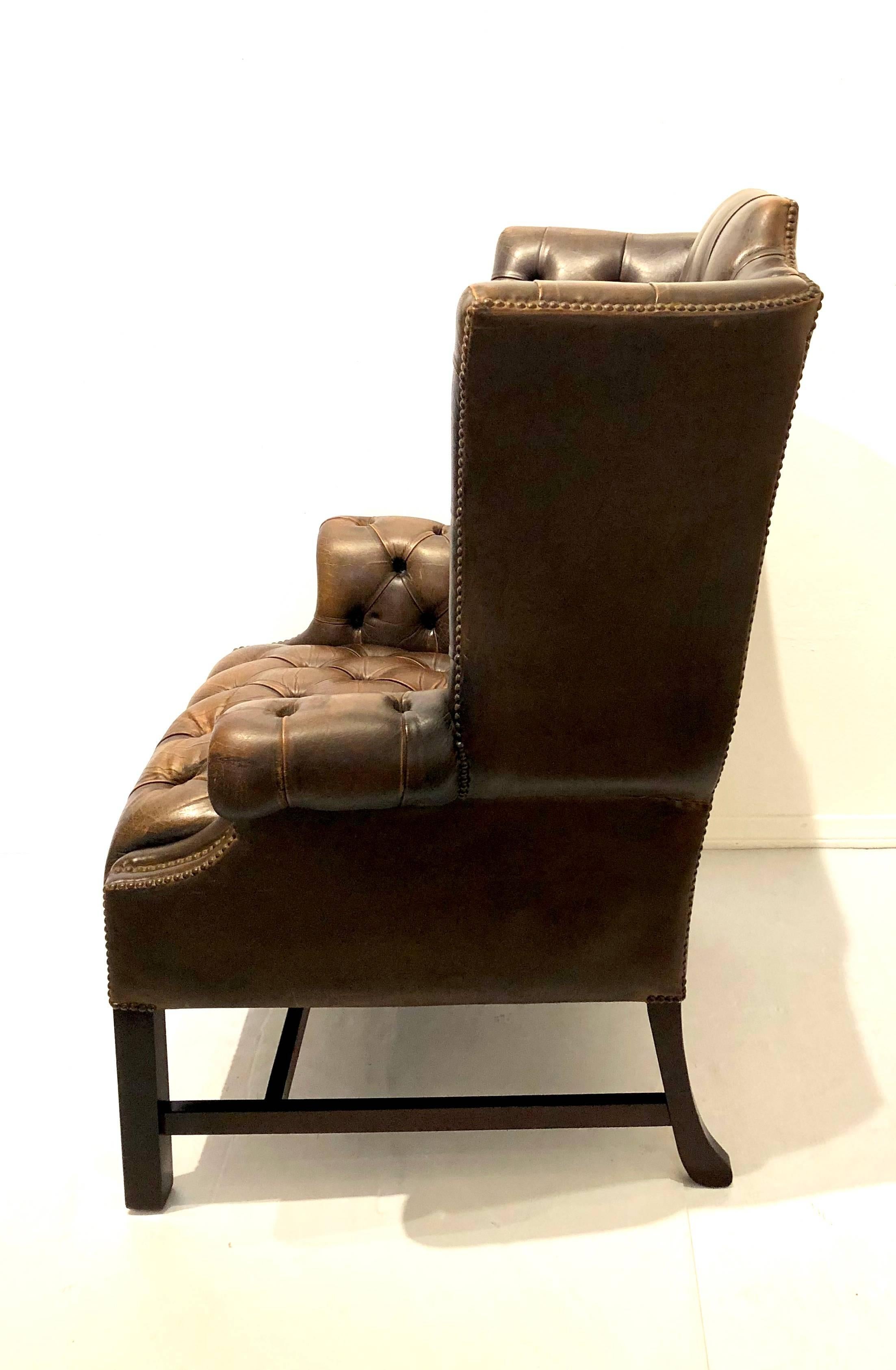 A classic antique English Chesterfield wing-back chair upholstered in brown distressed brown leather with tufted back and seat decorative nail heads around the silhouette. Chair base on square Marlboro front legs and splayed back legs that are