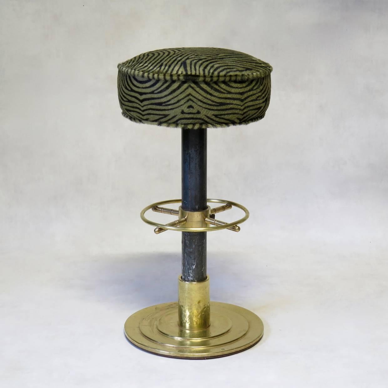 Nice-quality stool, with brass details. The stepped base is made of hammered brass, and the foot rail, fashioned like a boat wheel, is solid brass. The seat is upholstered in faux-zebra calf hair. From a boat. Very heavy-based.