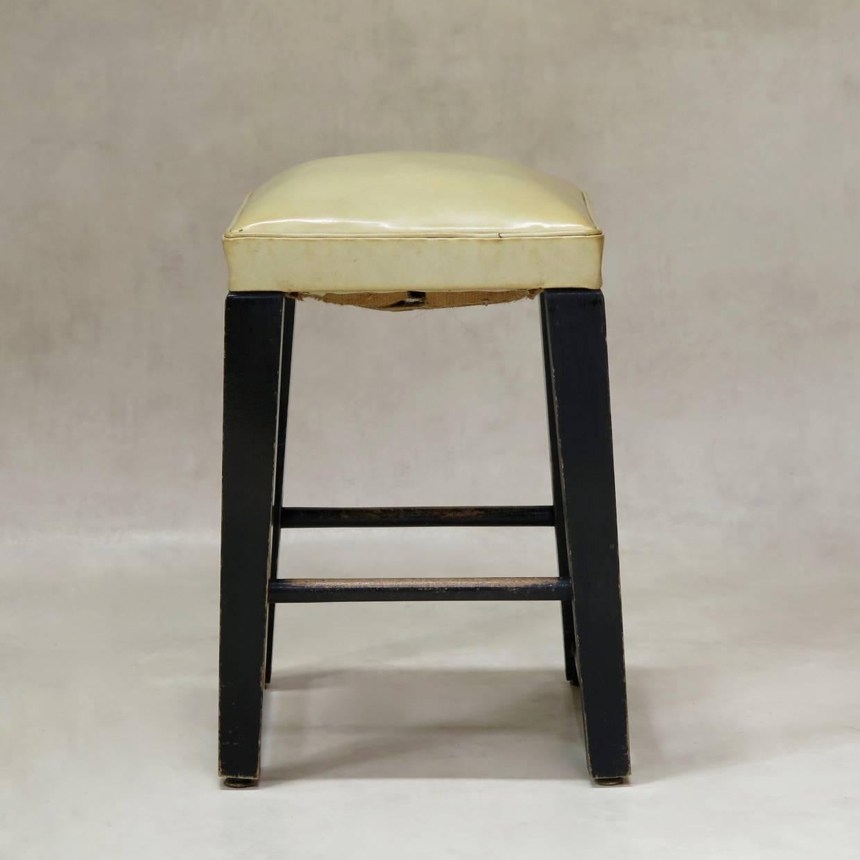 Striking pair of modernist stools. The bases are painted black (now rather worn) with tapering, block legs, slightly splayed and joined together by foot rests on two different levels. The seats are covered with the original ivory-coloured naugahyde