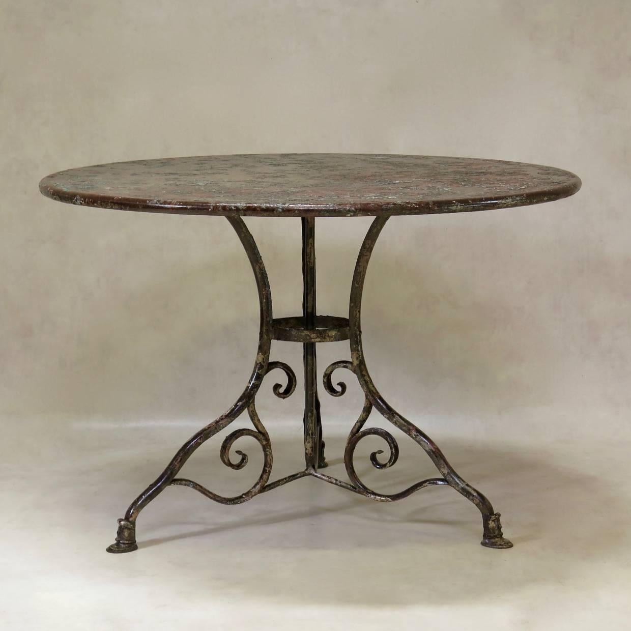 Rare and elegant round outdoor dining table raised on a very pretty tripod base, ending in sabots. The table is made of solid, heavy wrought iron and possesses an unusual palimpsest of muted colours (the dark grey of the iron, as well as dark green