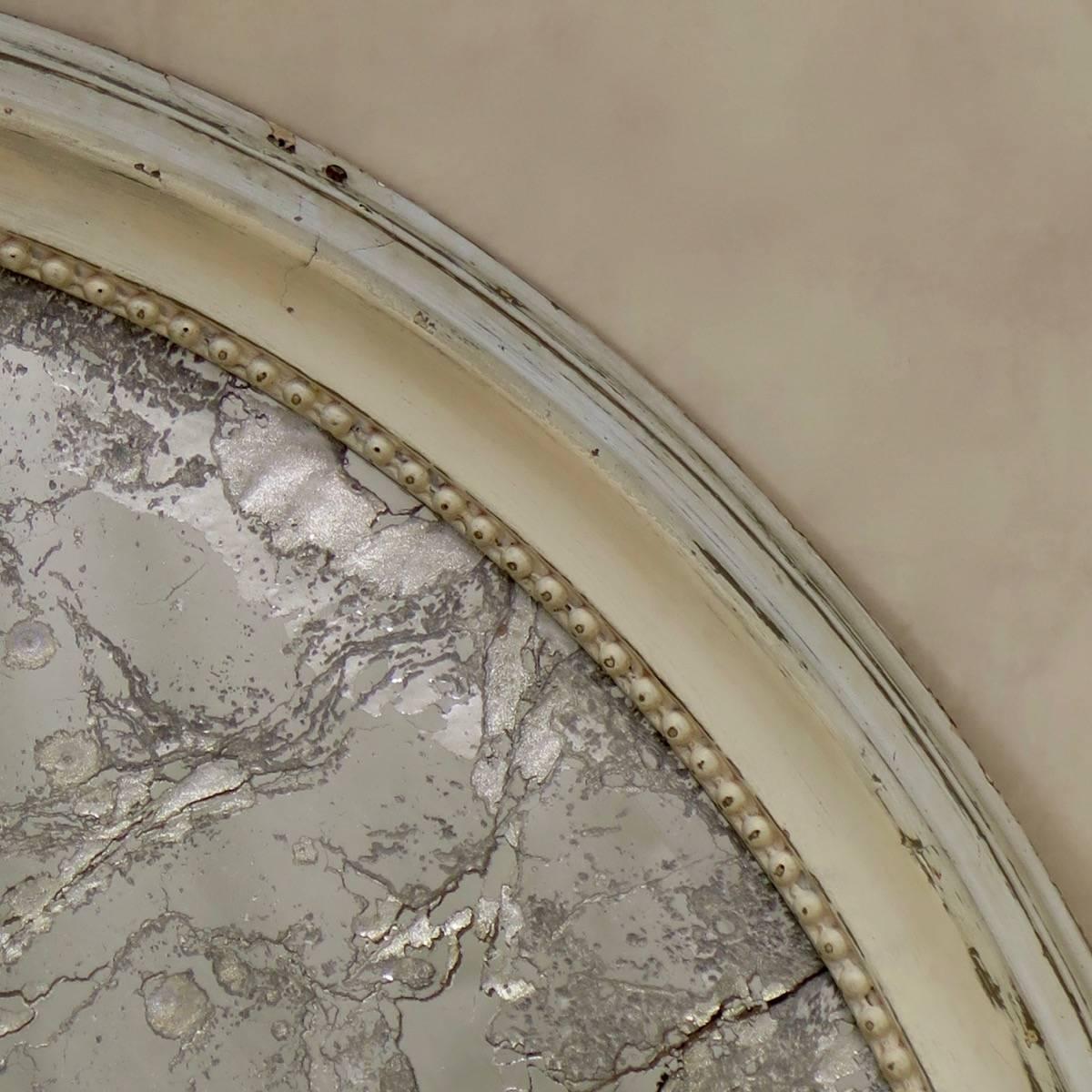 Elégant miroir ovale monté dans un cadre en bois moulé avec une garniture intérieure perlée et peint en blanc cassé. Verre biseauté, avec un support en argent vieilli et craquelé.