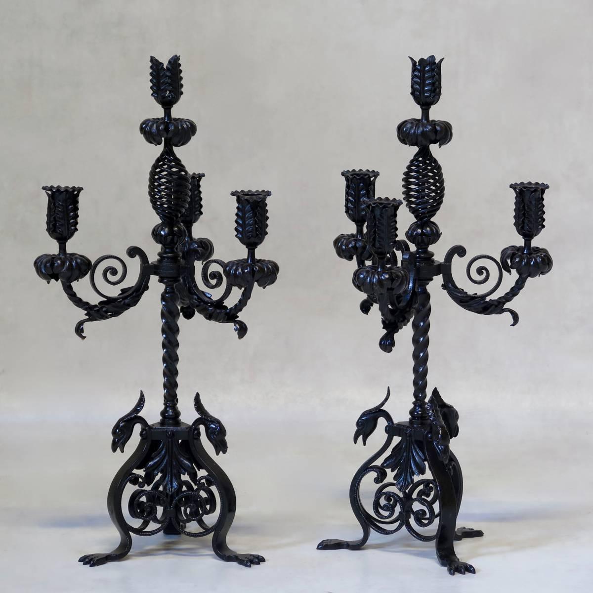 Pair of solid, wrought iron candleholders, with three arms each, decorated with scrolling acanthus leaves, raised on a twisted iron stem. The three-sided bases rest on claw feet and are mounted with phoenix heads at each corner. Beautiful