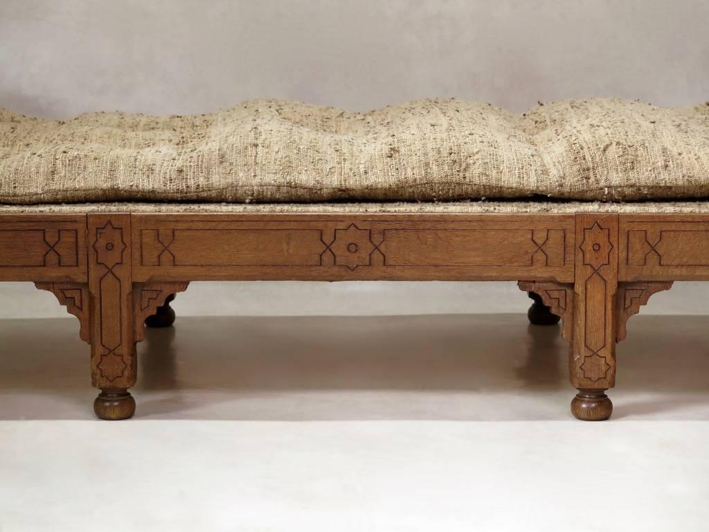 Exotic and elegant daybed in the oriental taste, with an oak base engraved with geometric motifs and raised on bun feet, covered in a marled beige wool fabric, with a thin padded mattress and a cushion.
