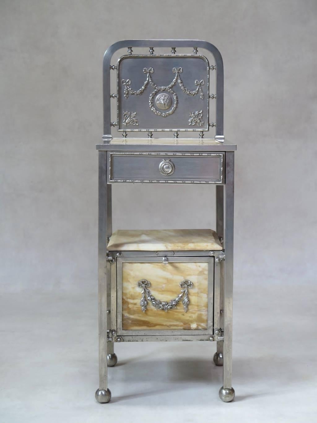 Sturdy brushed steel bedside table with a yellow marble top and cabinet, raised on chunky ball feet, and decorated with Louis XVI-style wreaths and ribbons.