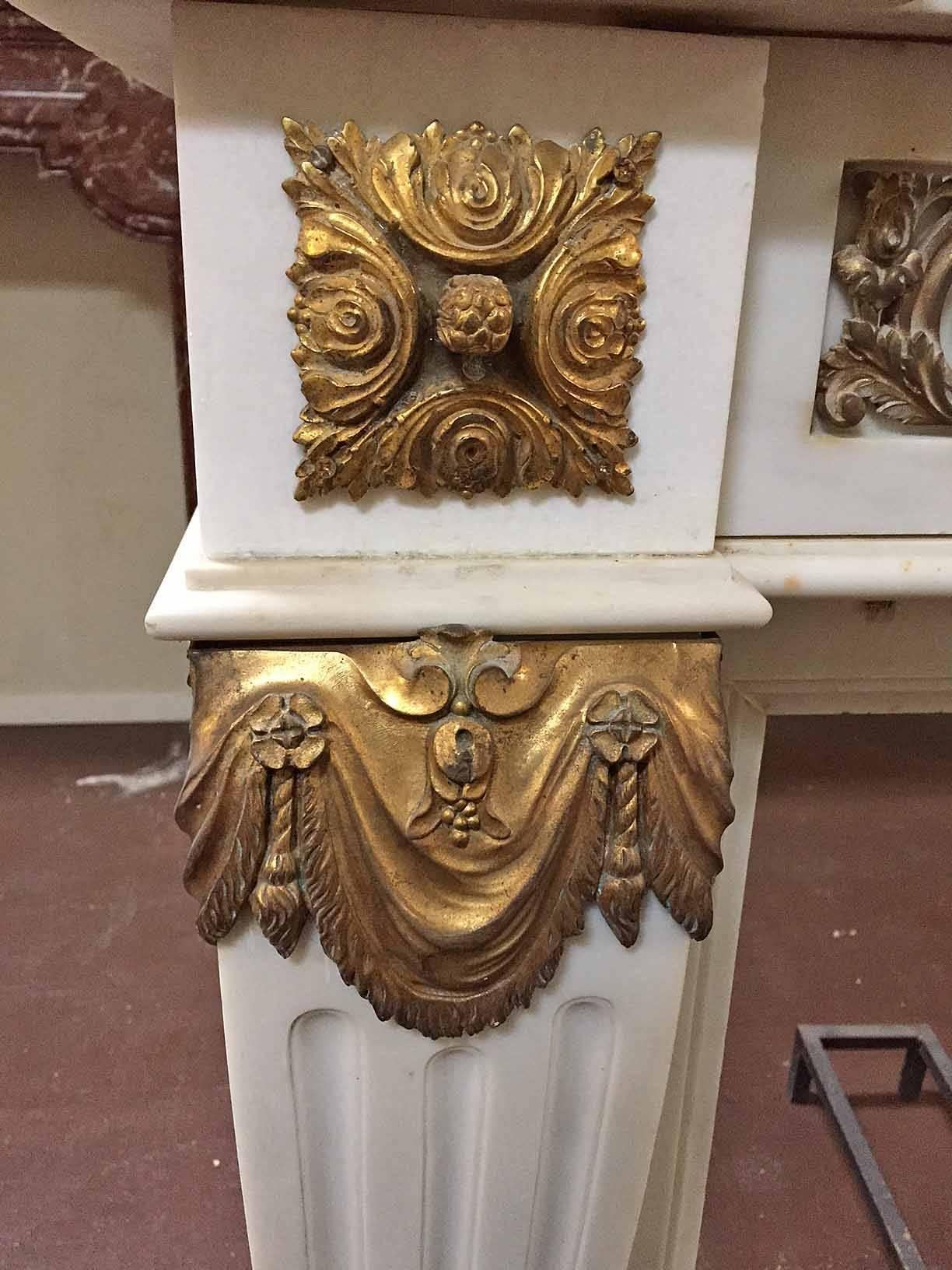 This French Carrara marble mantel bares bronze accents throughout the body. The bronze medallion reflects three cherubs playing as well as scroll-like designs across the lentil. The legs bare bronze drapes and reverse fluting. This is an exquisite