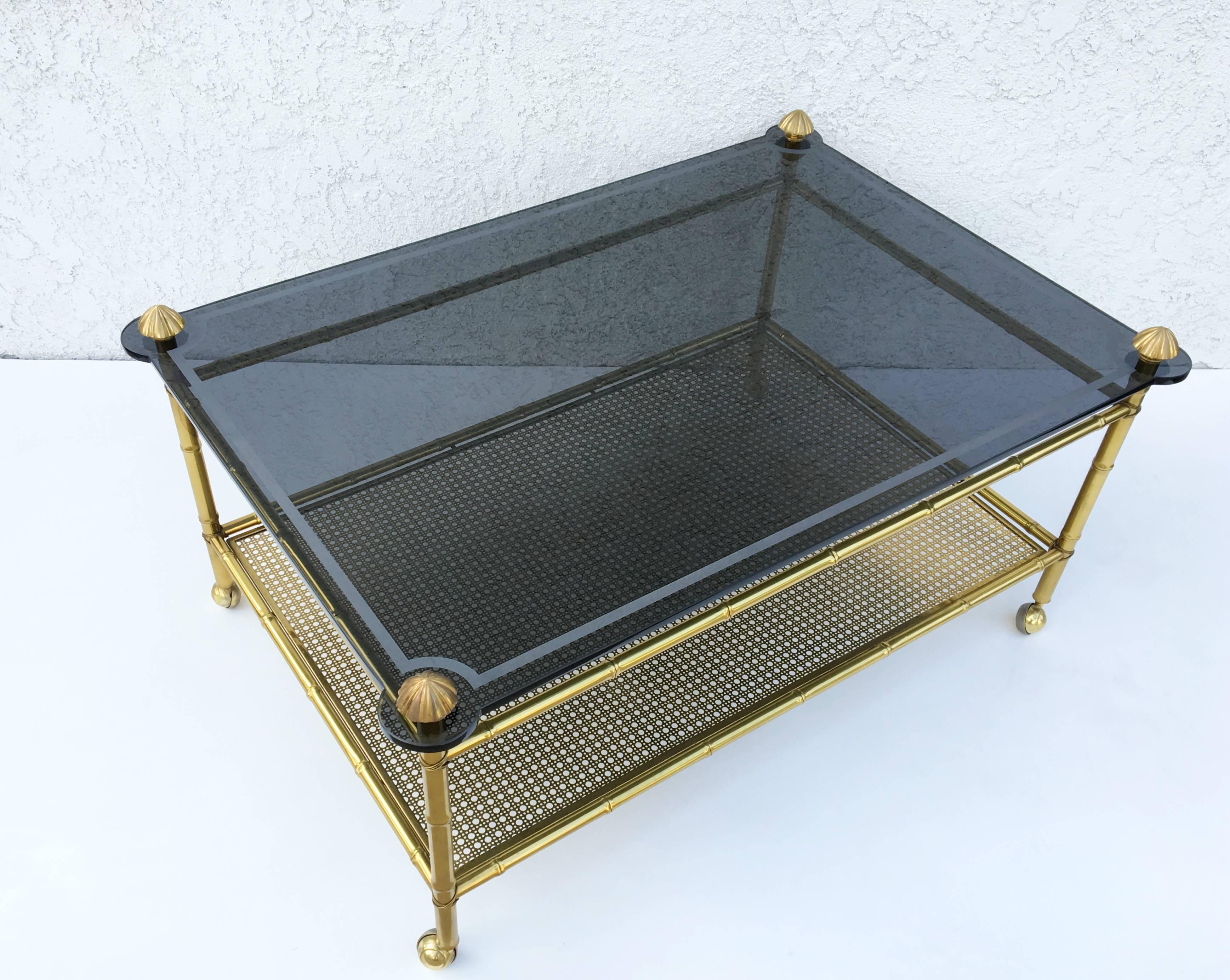 A glamorous 1960s French two-tiered brass faux bamboo and smoked glass top with a acidic edge design on casters. 
The glass is 1/2