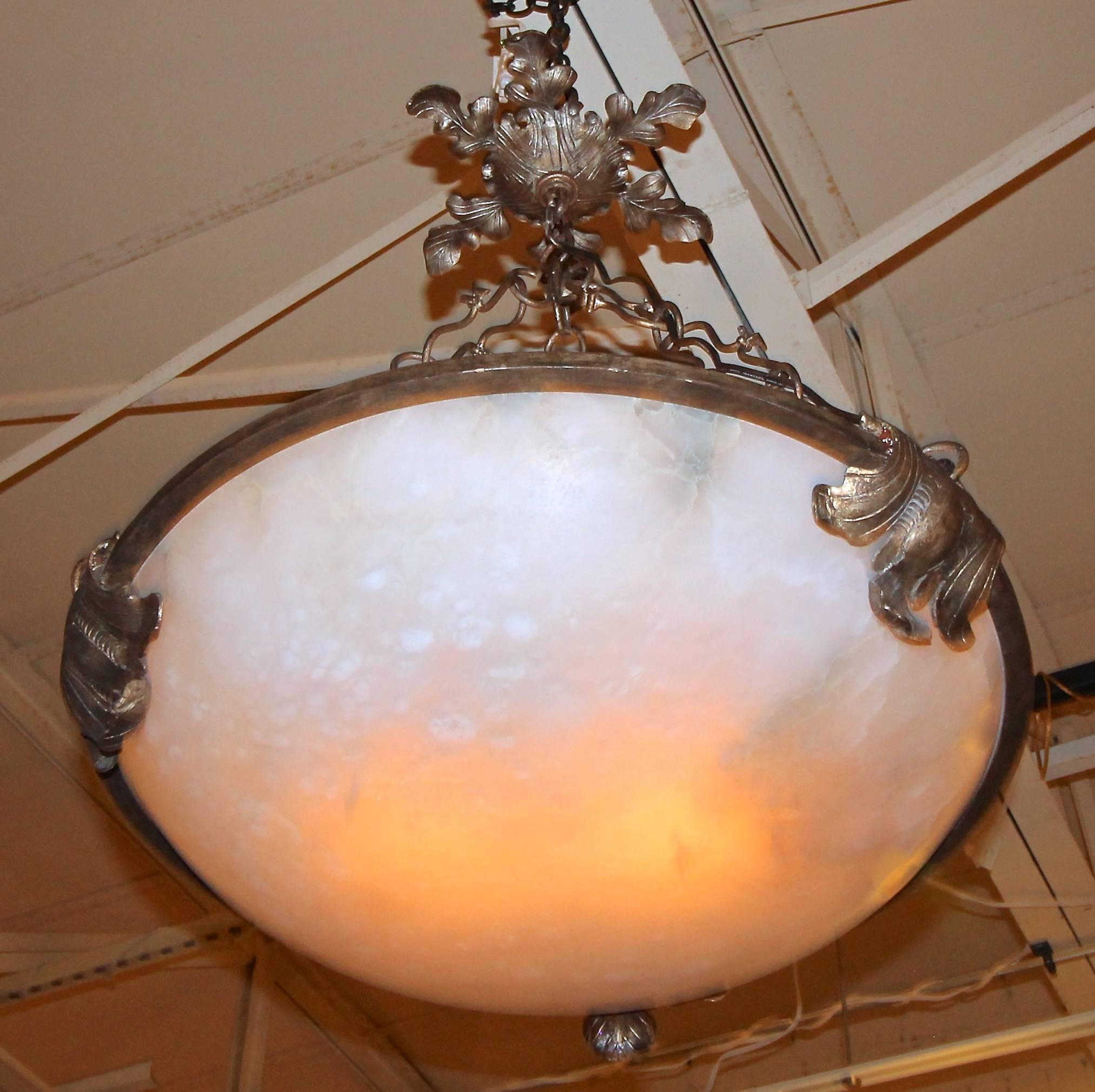 A dramatic pair of alabaster pendant light fixtures or chandeliers with hand-wrought acanthus scrolled ceiling cap and surround. Hand-forged chain links. Each light fixture uses 2 - 60 watt max "A"/Edison type bulbs. Large and dramatic