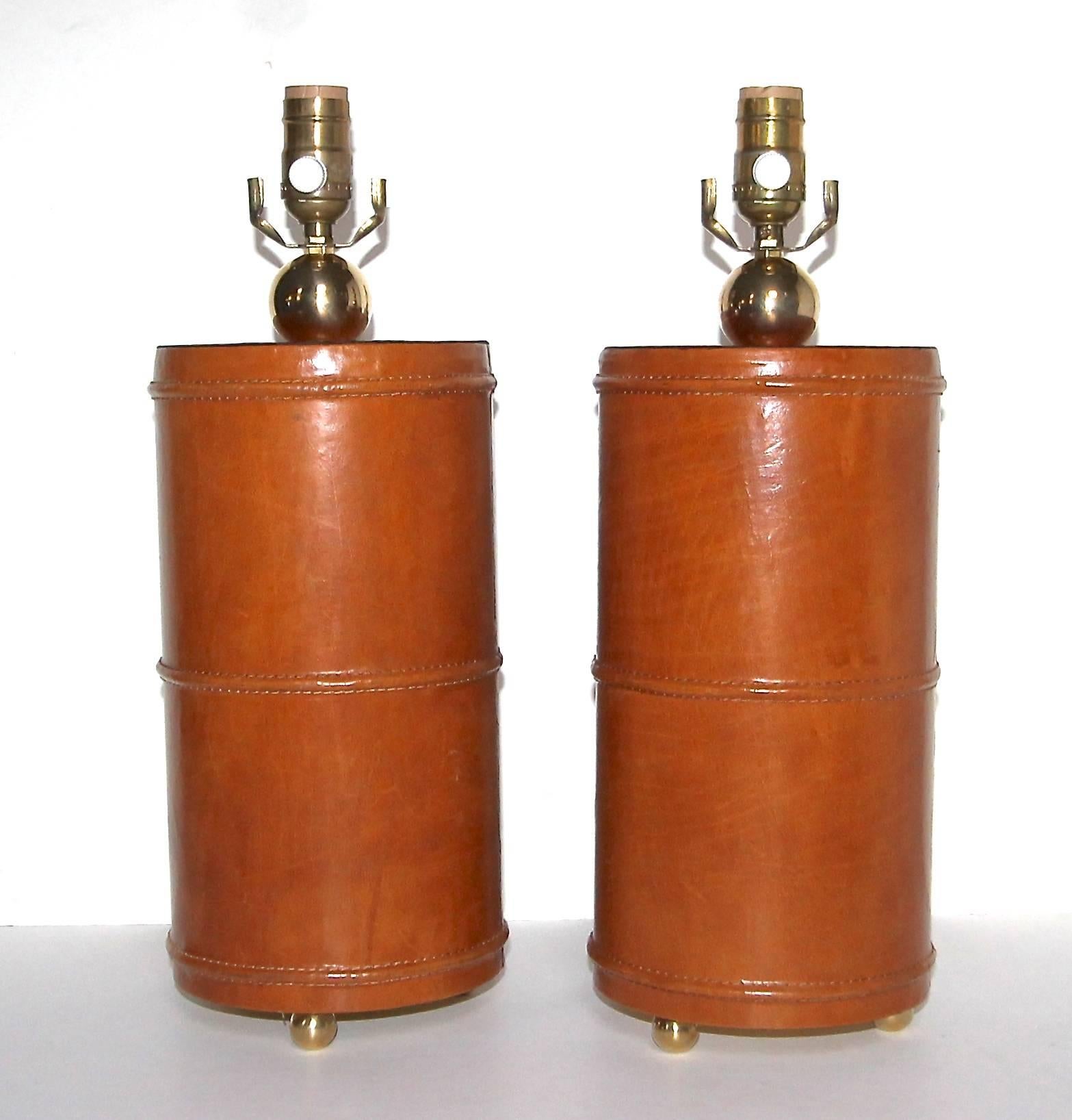A handsome pair of cylindrical leather wrapped table lamps with stitched detailing in the manner of Jacques Adnet. Base is raised on three brass ball feet with a large solid brass ball at the neck. Newly wired for the US with French style rayon