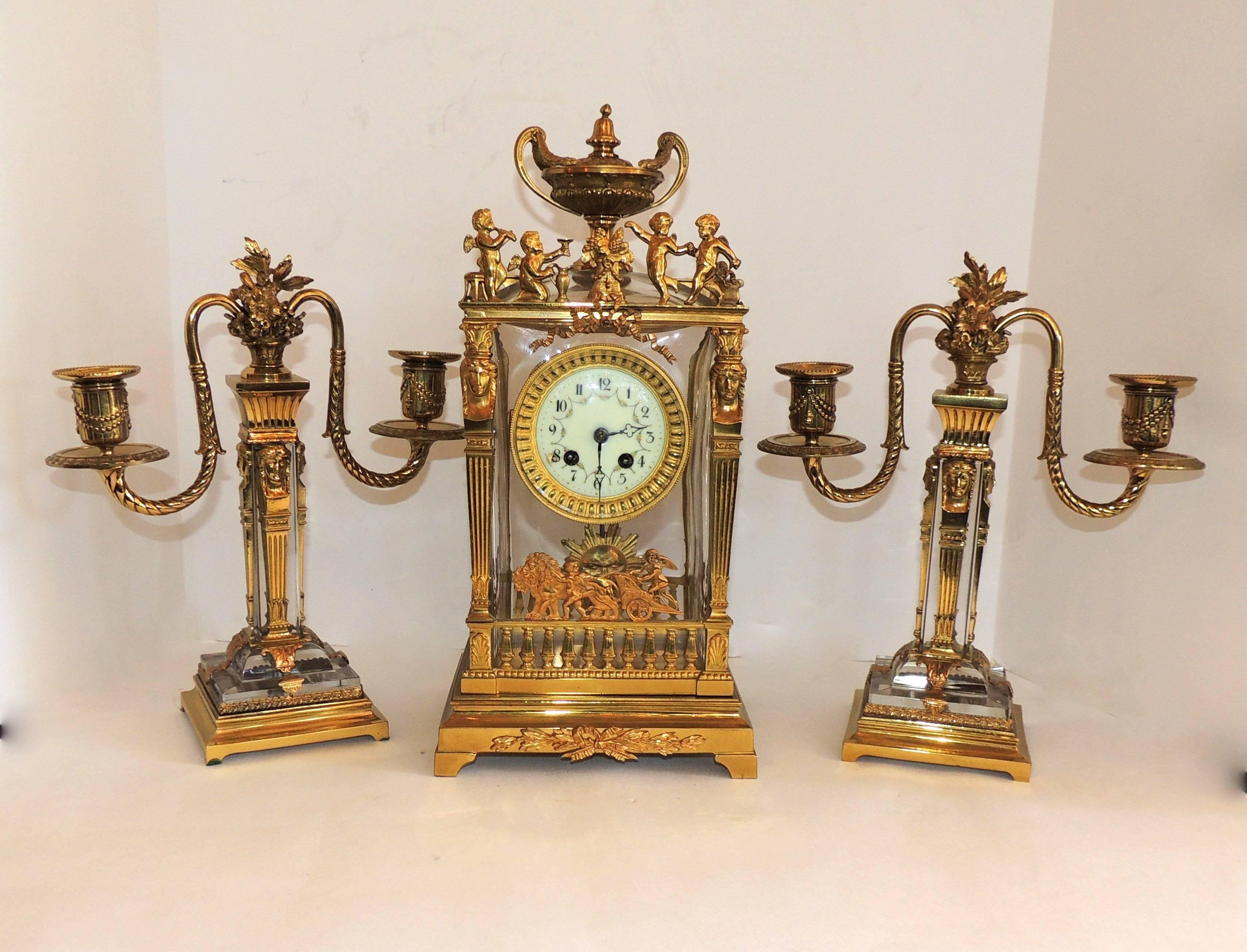 A wonderful Empire crystal clock with lots of details in the Dore Bronze of this three-piece clock set. The clock is surrounded by cherubs on both the top and bottom, with a smiling sun on the pendulum. Fluted detail is on both the candelabras and