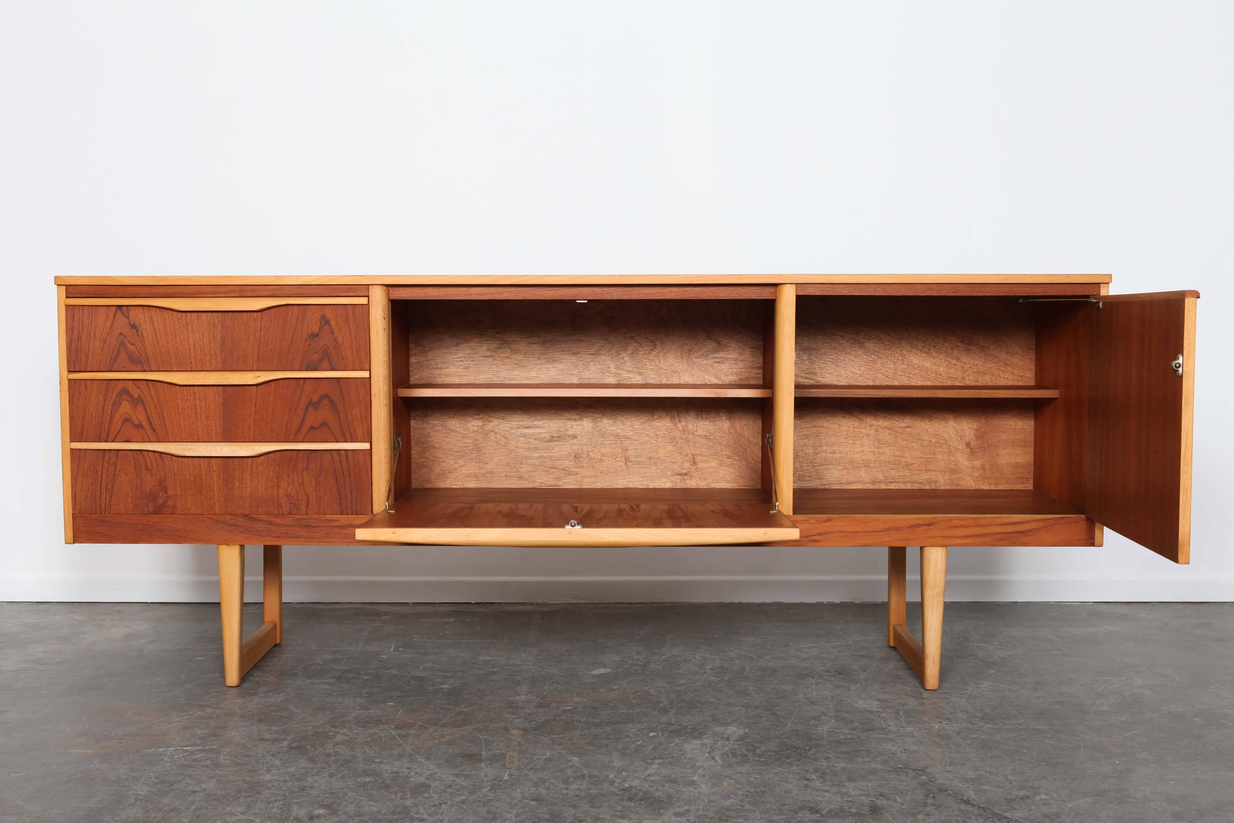 English Mid-Century Modern teak two-door, three drawer low sideboard with contrasting light handles, trim and sleigh style legs with matching cathedral grain running throughout the front. Newly refinished in a natural teak oil finish.