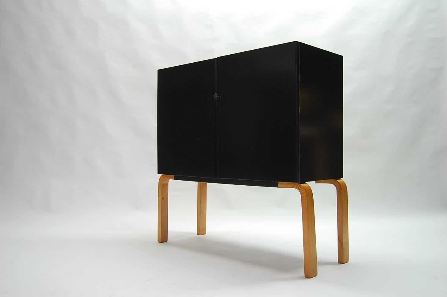 Alvar Aalto two door cabinet with adjustable shelves, circa 1938. Cabinet in new black lacquer, legs in natural birch. Doors are lockable, brass key and escutcheon. Interior has five adjustable shelves (two half depth shelves and three full depth