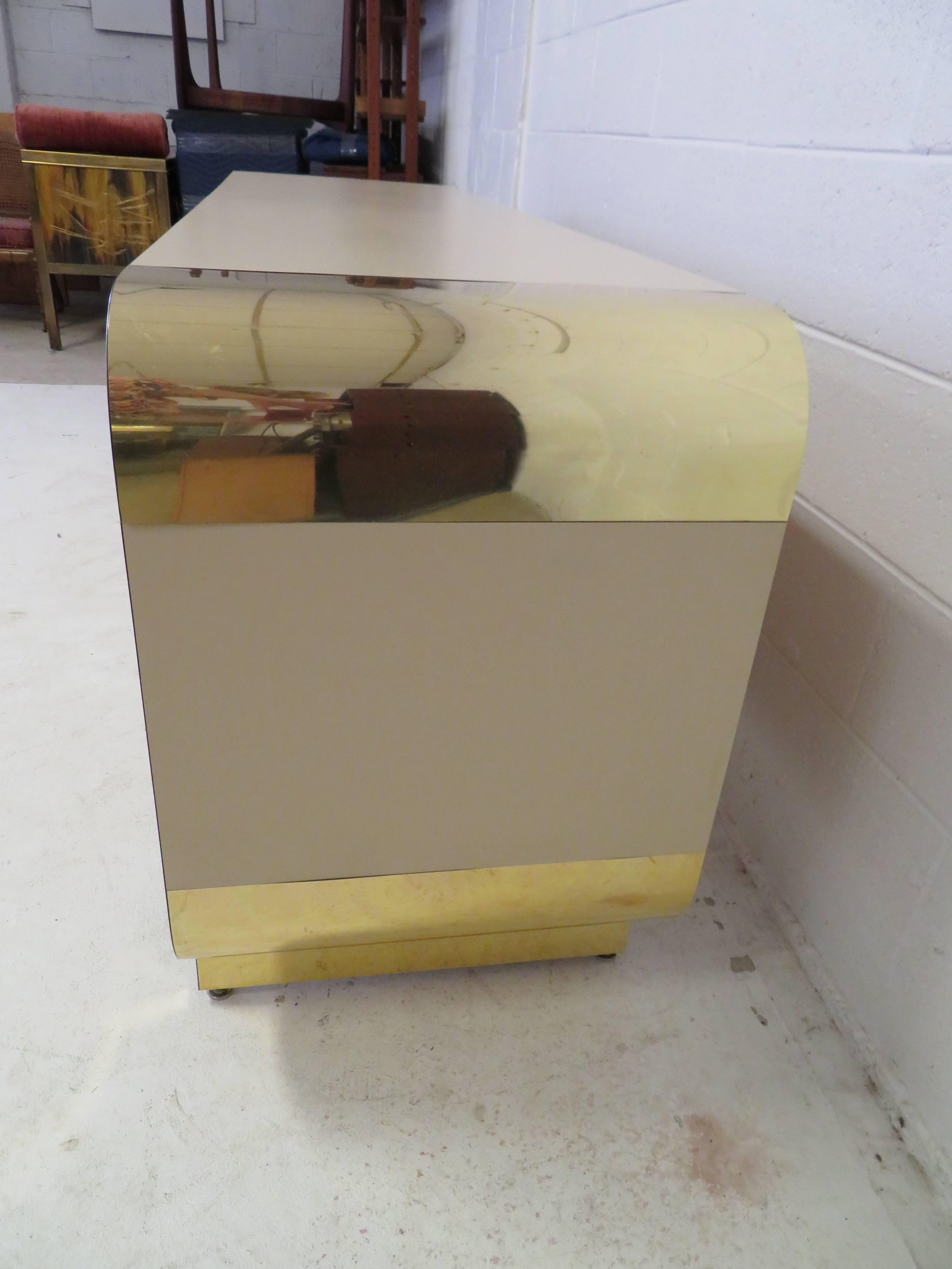 Insolita credenza con bordo curvo in ottone della collezione Pace Collection. Questo pezzo è impiallacciato con un laminato color tortora per una facile manutenzione e presenta eleganti bordi e piedini curvi in ottone. Le porte si aprono per