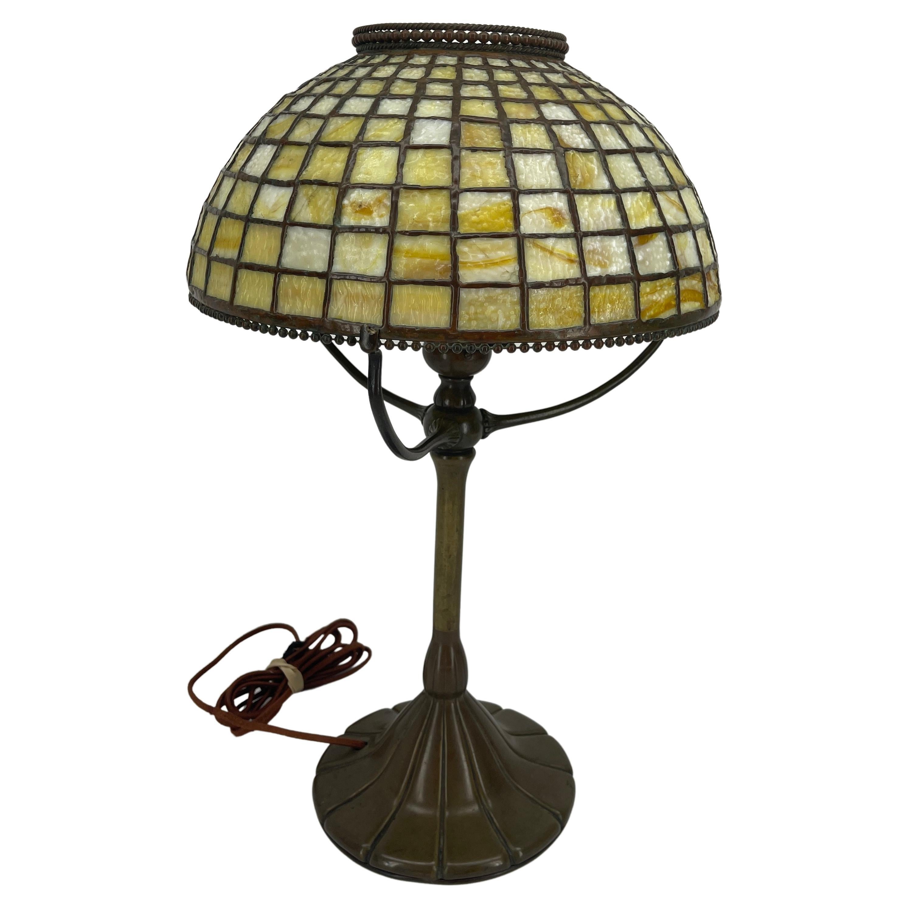 This vintage early 20th Century leaded glass and bronze table lamp was made by the Tiffany Studios, New York. The handcrafted leaded shade is made up of yellow/amber stained tiles and features a beautiful bronze fringe. The shade rests upon a tripod