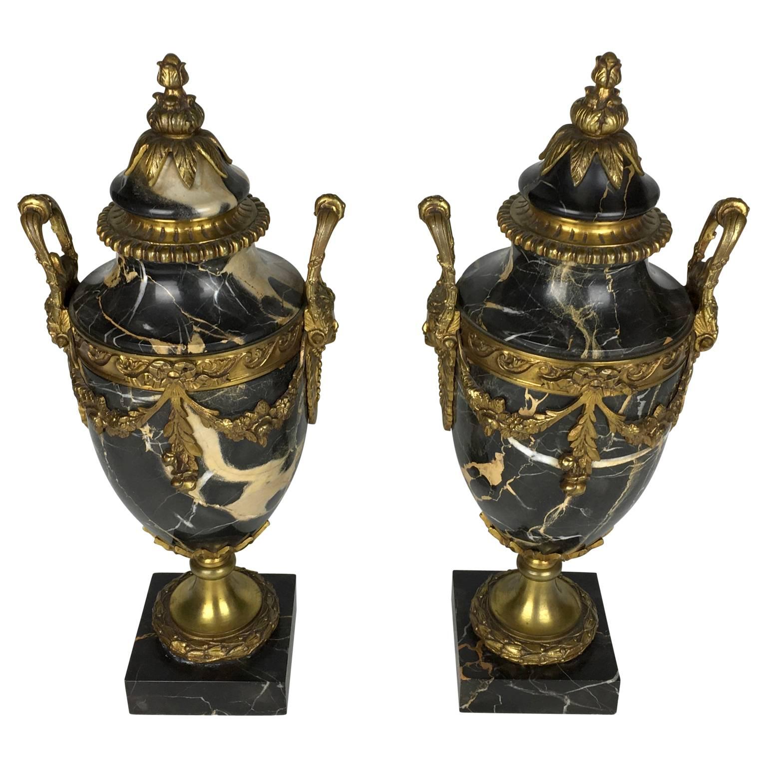 A large impressive pair of French Louis XVI style black marble urns with gilt bronze ormolu mounts. Draping floral swag. Laurel leaf design, face designed handles. Beautiful veining to the marble.