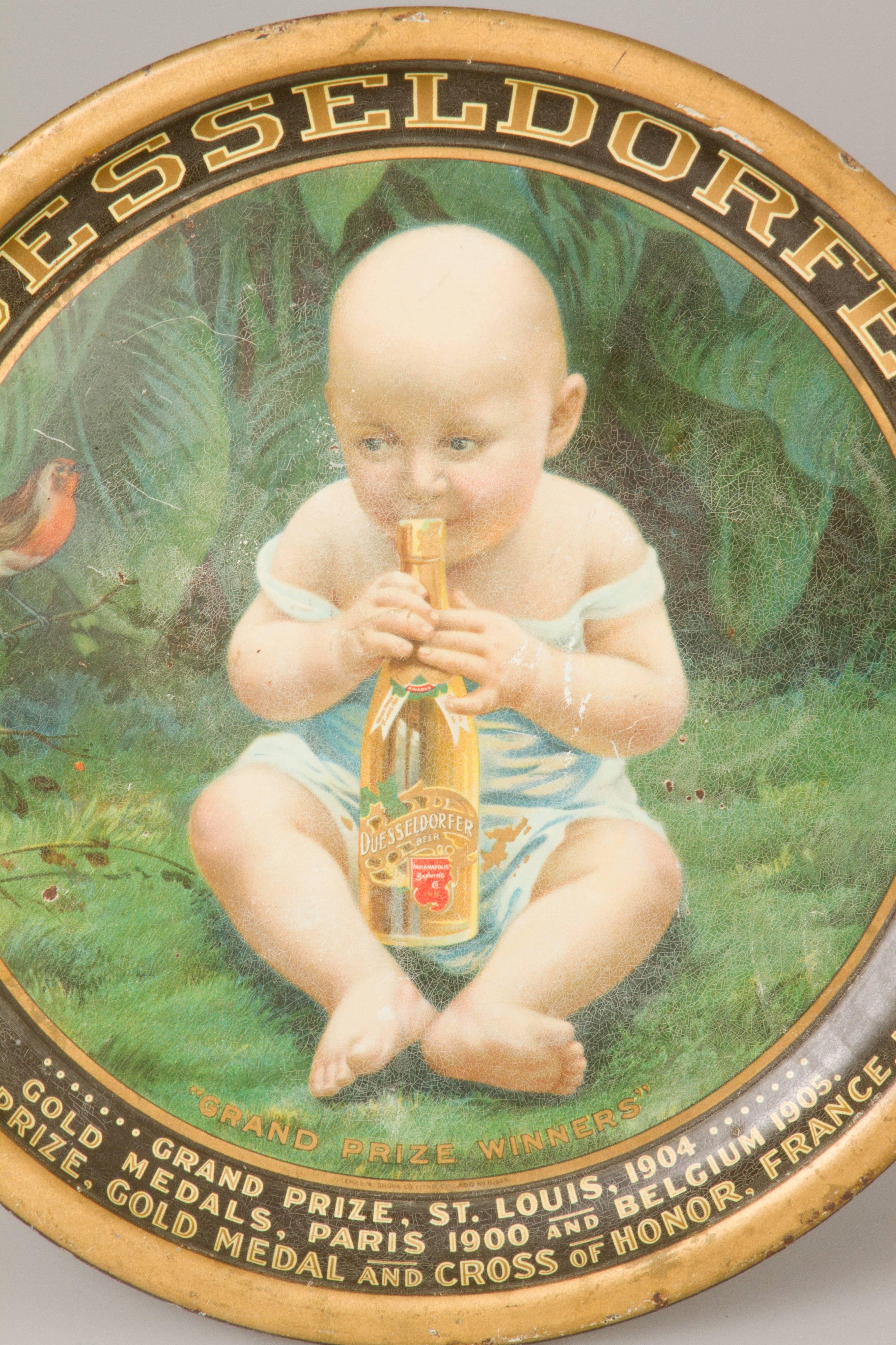 This beer serving tray for Duesseldorfer Beer shows a baby biting the top of one of a beer bottle. This tray lists the medals this company has won along the bottom edge: Grand prize, St. Louis, 1904. Gold medals, Paris, 1900 and Belgium, 1905. Grand