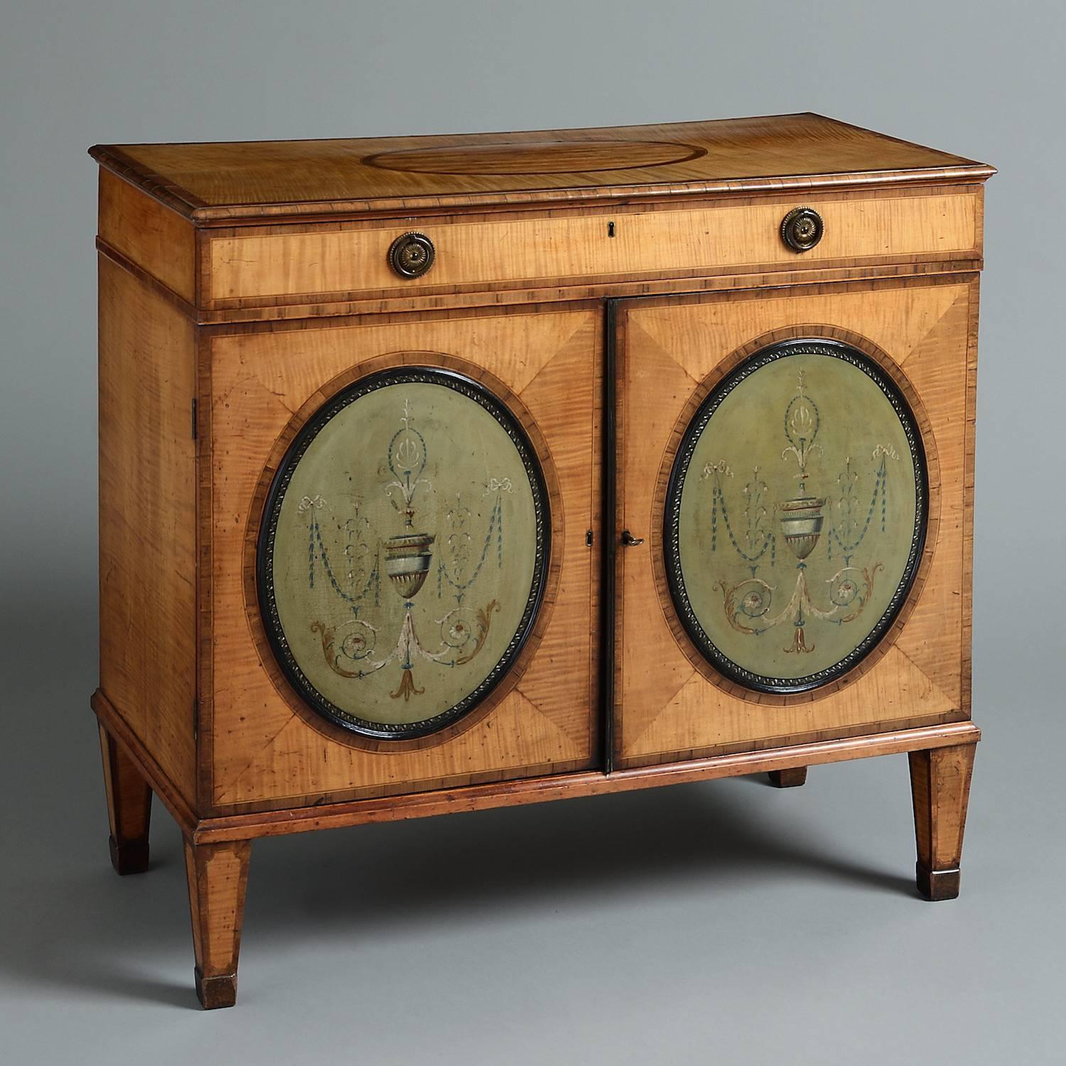 A rare George III satinwood side cabinet inset with painted tôle panels, circa 1790.

Banded in kingwood, the mahogany-lined frieze drawer with original gilt brass handle, the doors inset with convex oval tole panels painted with urns flanked by
