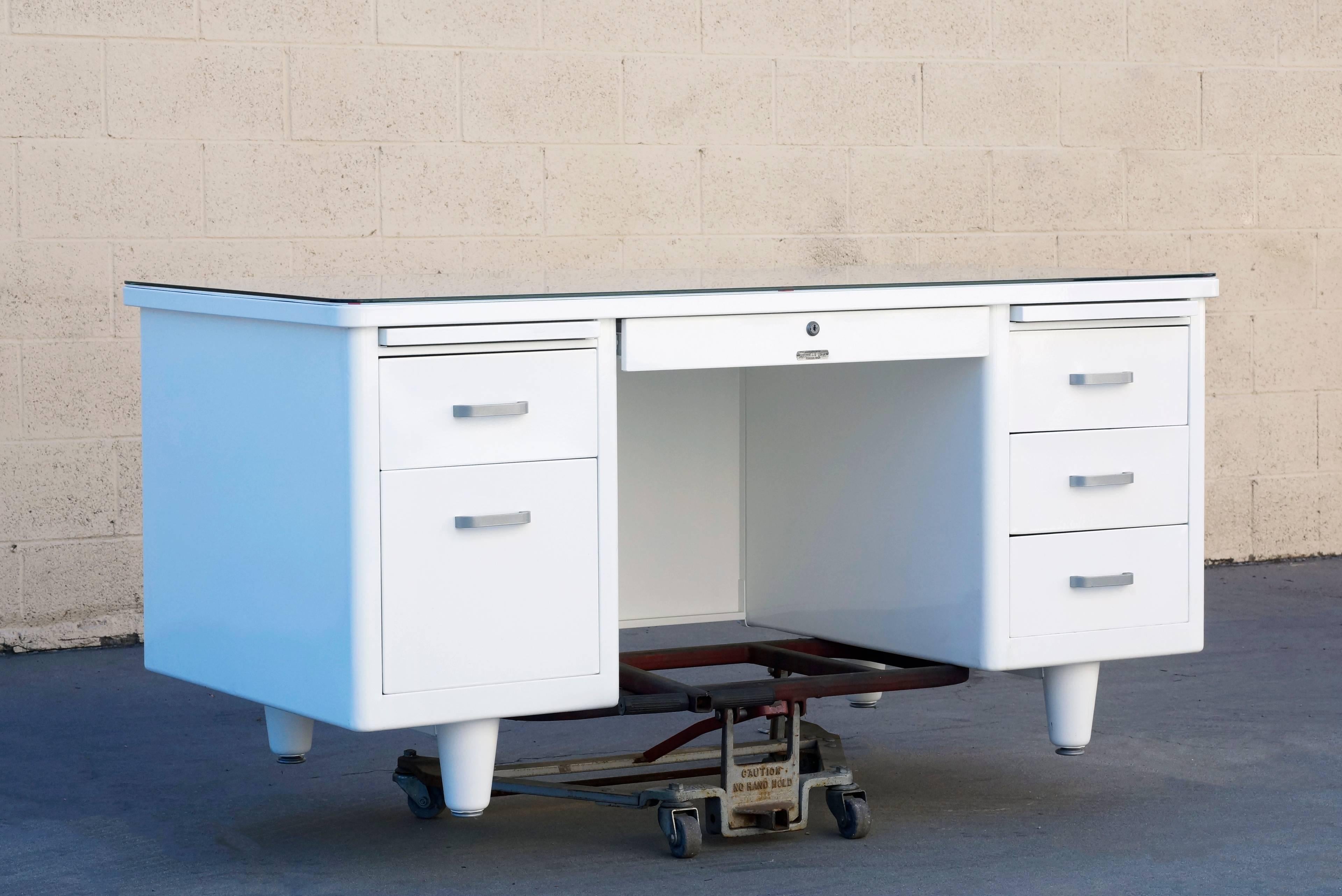 1960s McDowell Craig tanker refinished in gloss white with custom glass tabletop. This Classic double pedestal desk features many utility drawers, one filing drawer, one locking stationary drawer and two letter trays. All original aluminum hardware,