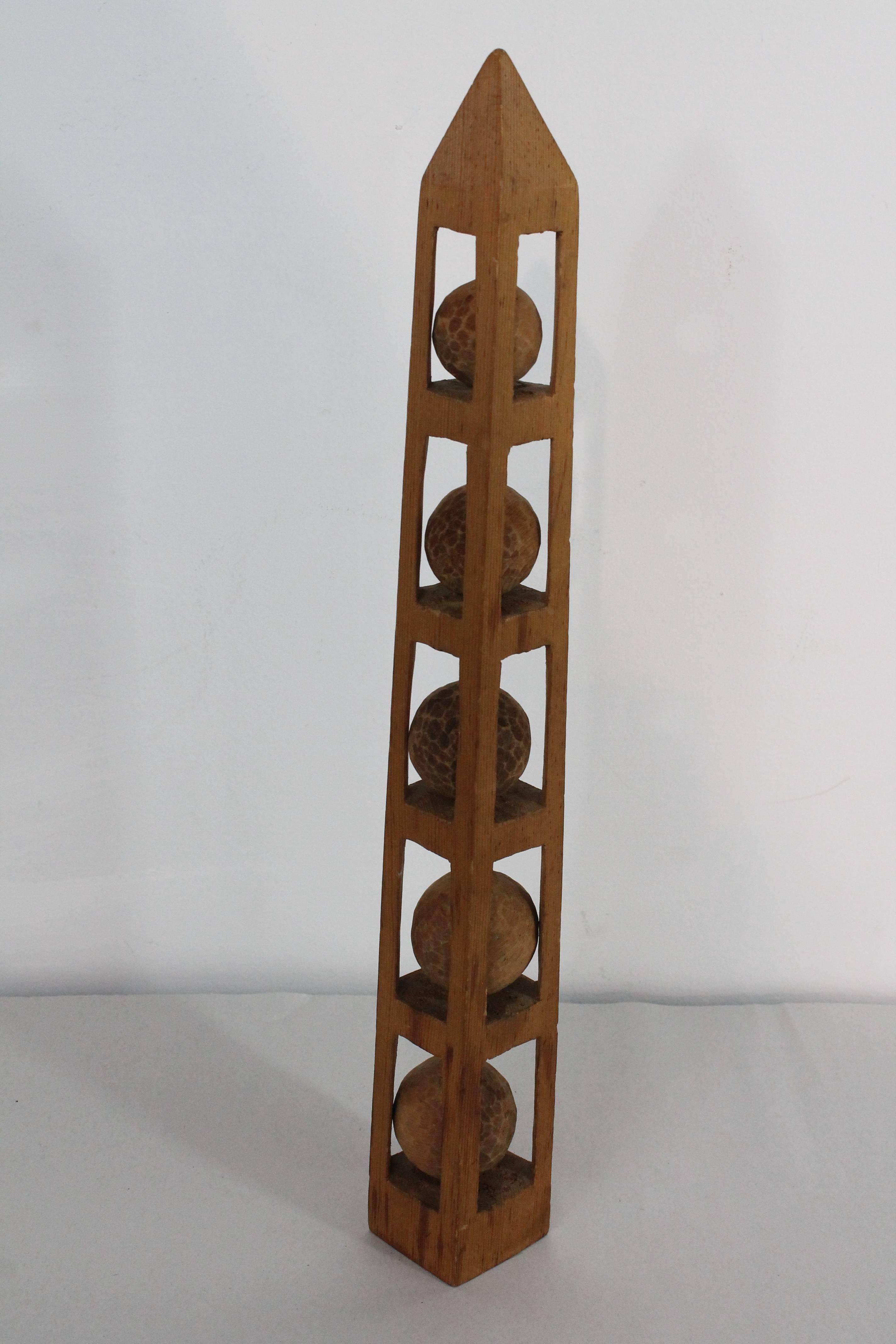 Folk art carved obelisk with 5 balls carved within the form. 
This whimsy shows the carvers skill by the balls being carved within the structure of obelisk form.