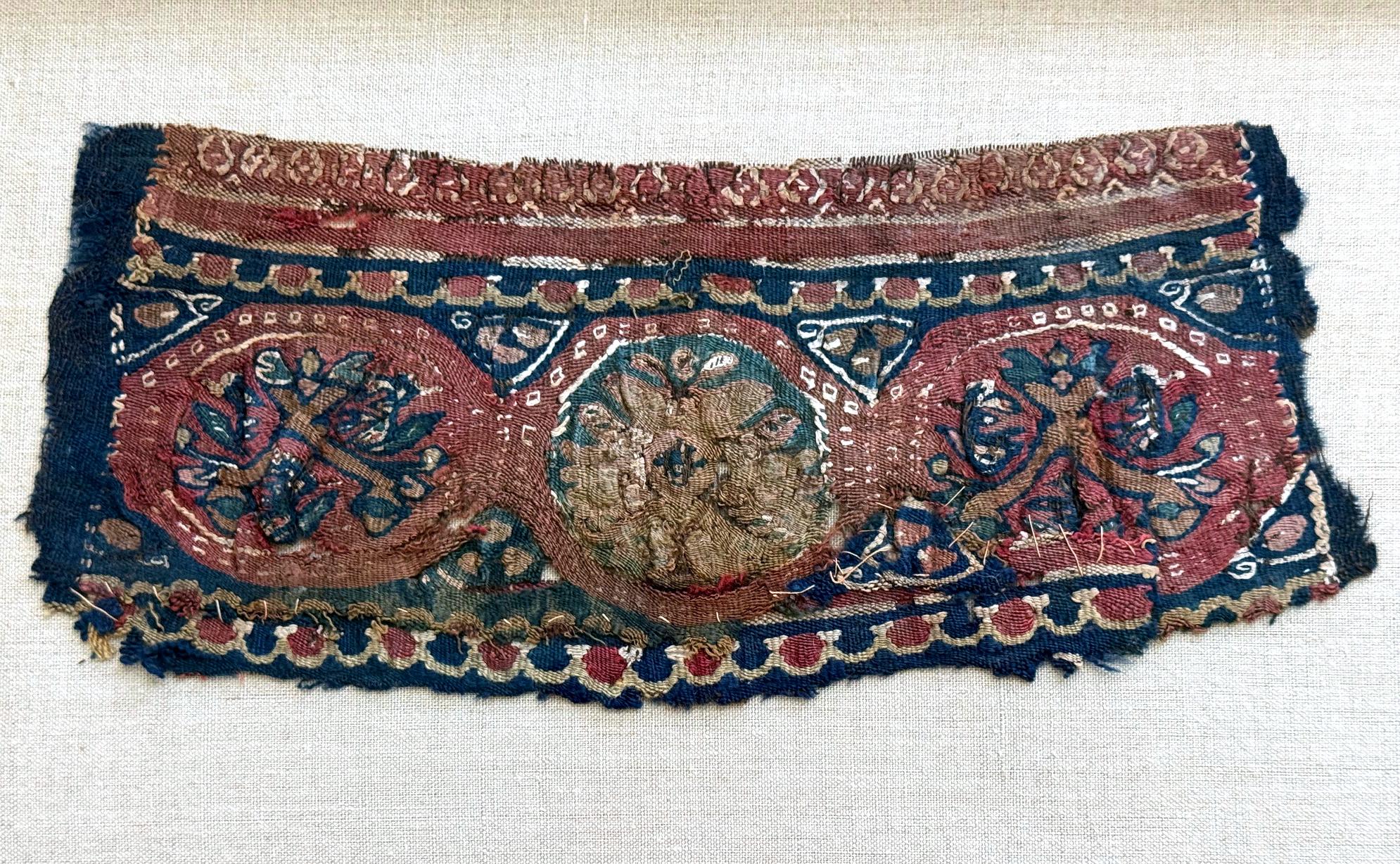 Ein kleines Stück eines antiken koptischen Textils, gewebt als horizontales Band mit zwei linearen Muschelbordüren, die drei zentrale Medaillons flankieren. Jedes Medaillon hat ein konzentrisches Muster mit einem floralen Cluster-Motiv in der Mitte.
