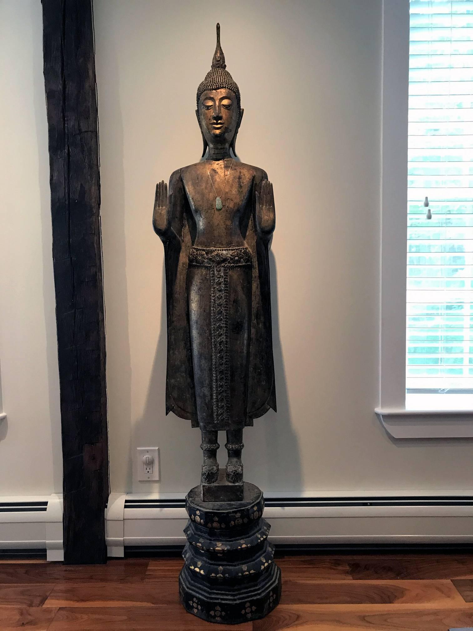 This wood Buddha in standing position was executed in a typical Laotian style, which features a slim and elongated body and extremely serene facial expression. His hands are in the gesture of double Abhaya mudra. This mudra symbolizes the act of