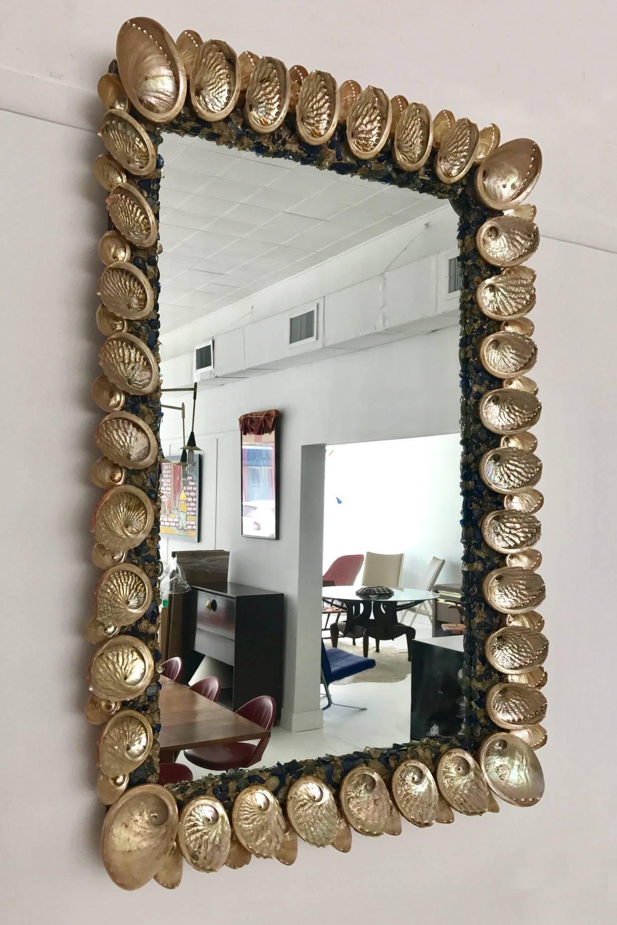 A beautiful handcrafted wall mirror in the style of Tony Duquette circa 1950-60s. It features an inner frame with mother-of-pearl finishes inset with blue color and clear glass pebbles, and an outer frame with abalone shells of alternating sizes.