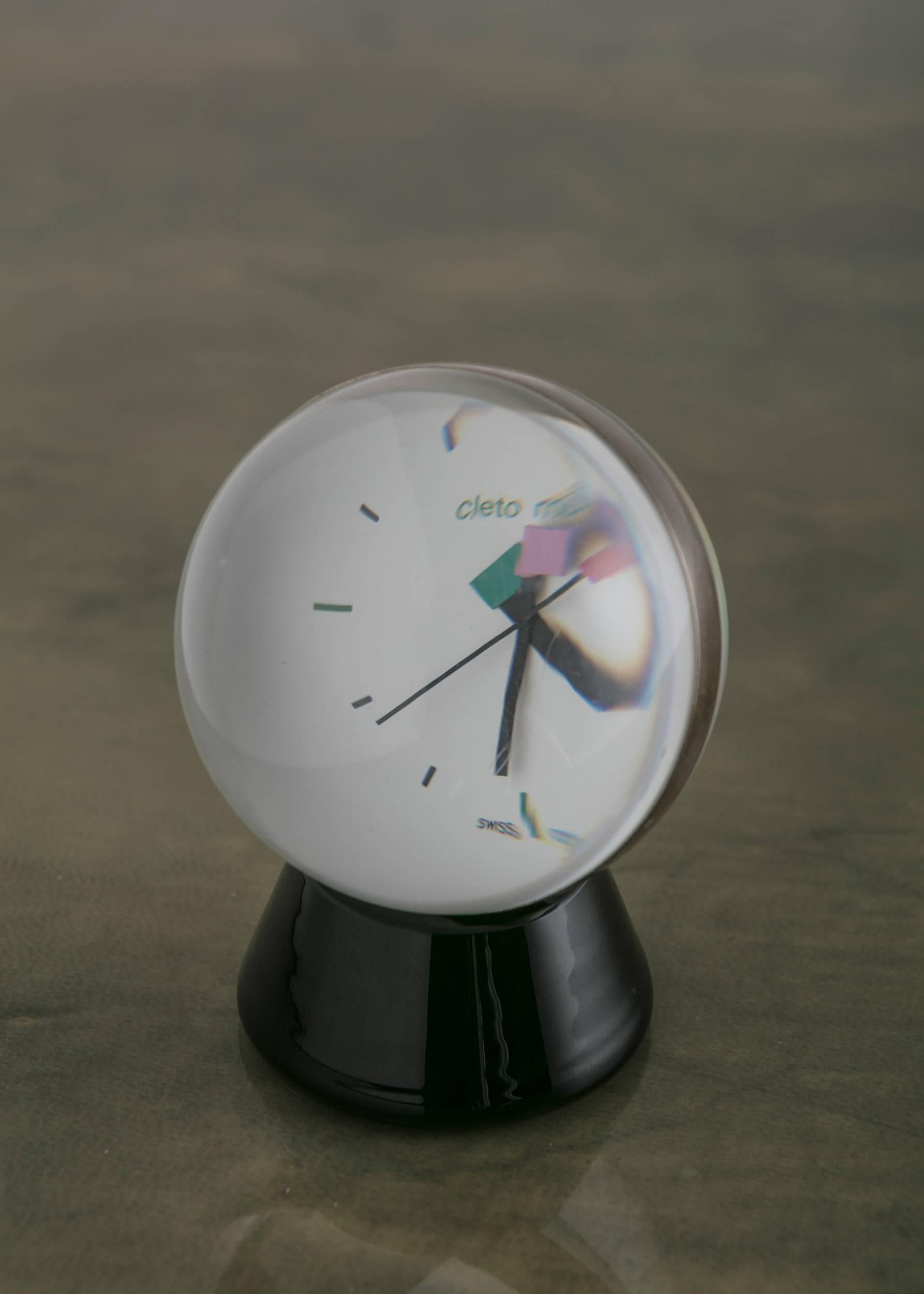 Minuscule horloge de table en verre de Cleto Munari.
Une lourde sphère transparente avec une bande argentée est posée sur un socle en verre noir.