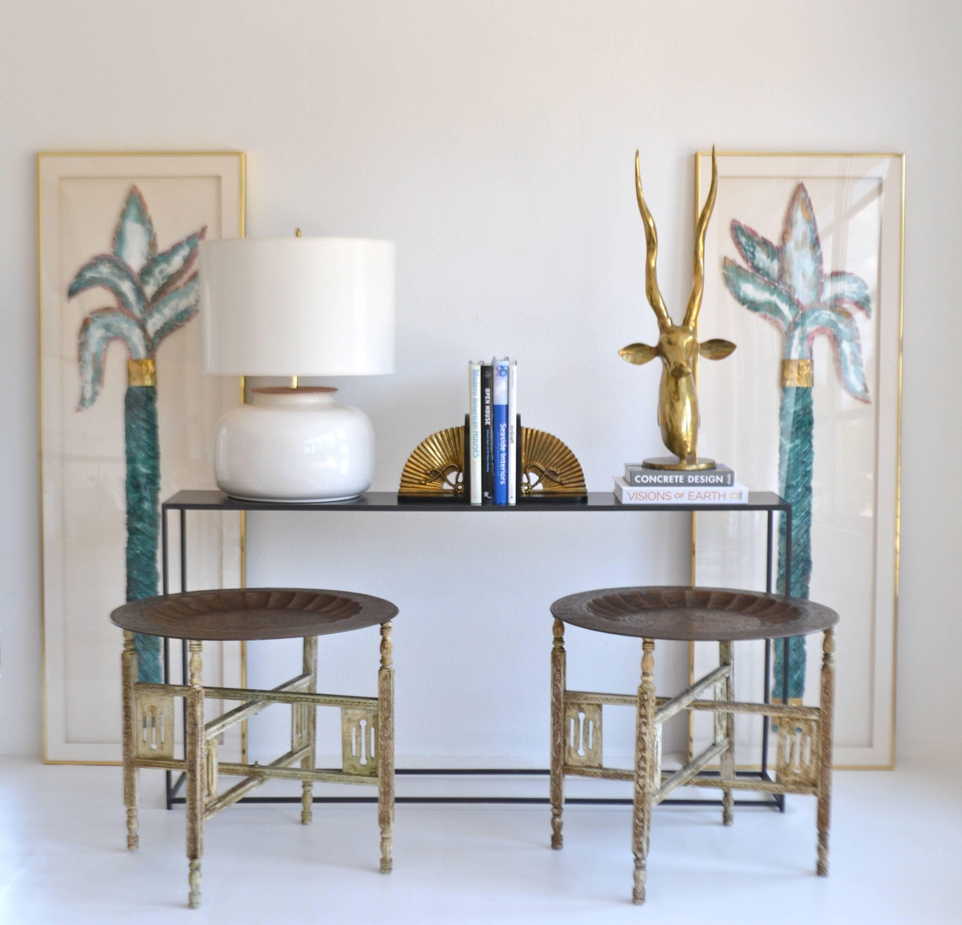 Striking pair of Anglo-Indian brass tray tables, circa 1940s -1950s. These sculptural side tables are designed of etched brass trays on distressed painted hand-carved wooden folding bases. The pair can also be versatilely used as a cocktail or