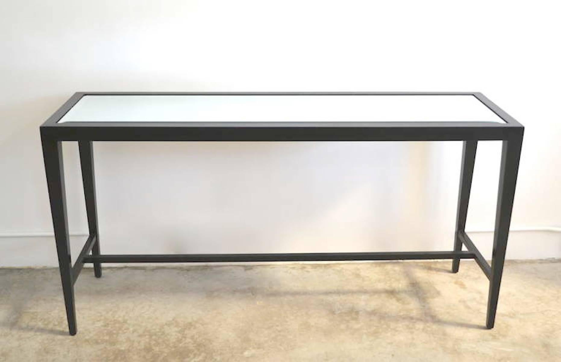 Stunning midcentury console table, circa 1950s-1960s. This striking, highly decorative sofa table is designed with an inset beveled mirror top and a polished black lacquered finish over its hardwood frame. The tapered design of the legs creates a