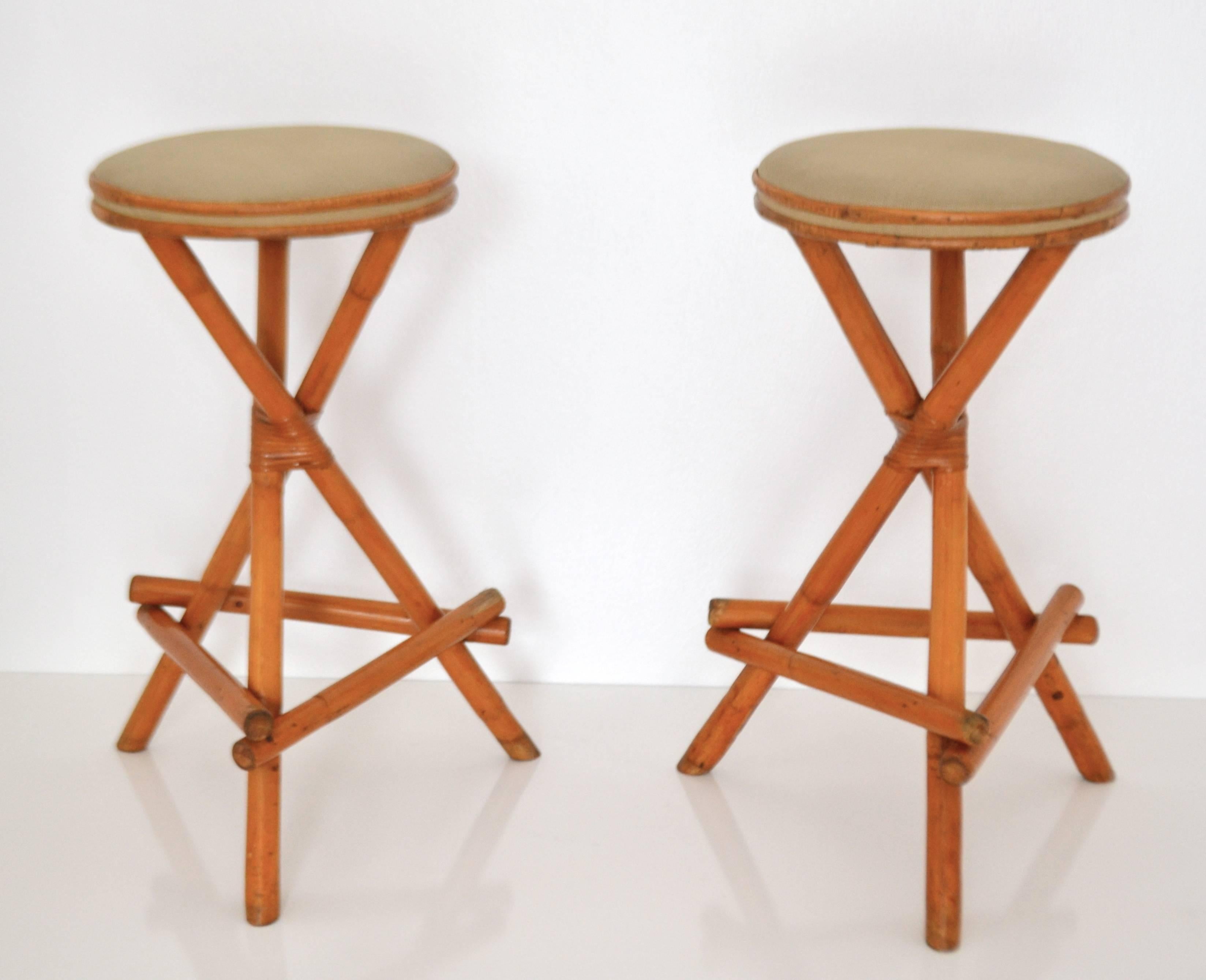 Striking pair of midcentury tripod form bamboo barstools, circa 1960s. These sculptural artisan crafted stools are designed of bent bamboo with cane wrapped accents and upholstered in a natural open weave canvas fabric.