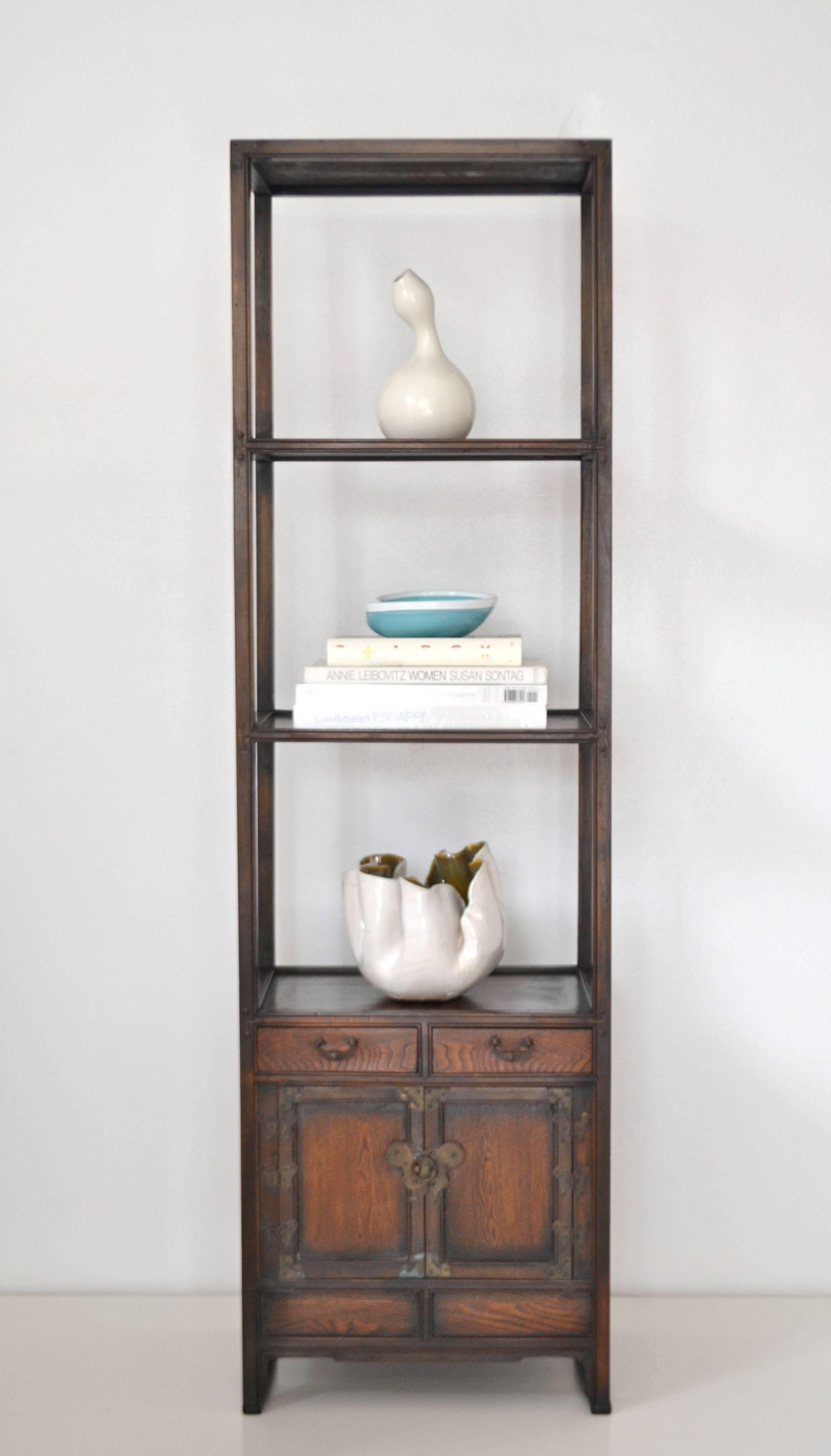 Stunning Asian elm bookcase, circa 1940s-1950s. This striking carved wood etagere is artisan crafted with three tiers of open shelves over two drawers and a double door cabinet accented with patinated bronze hardware.