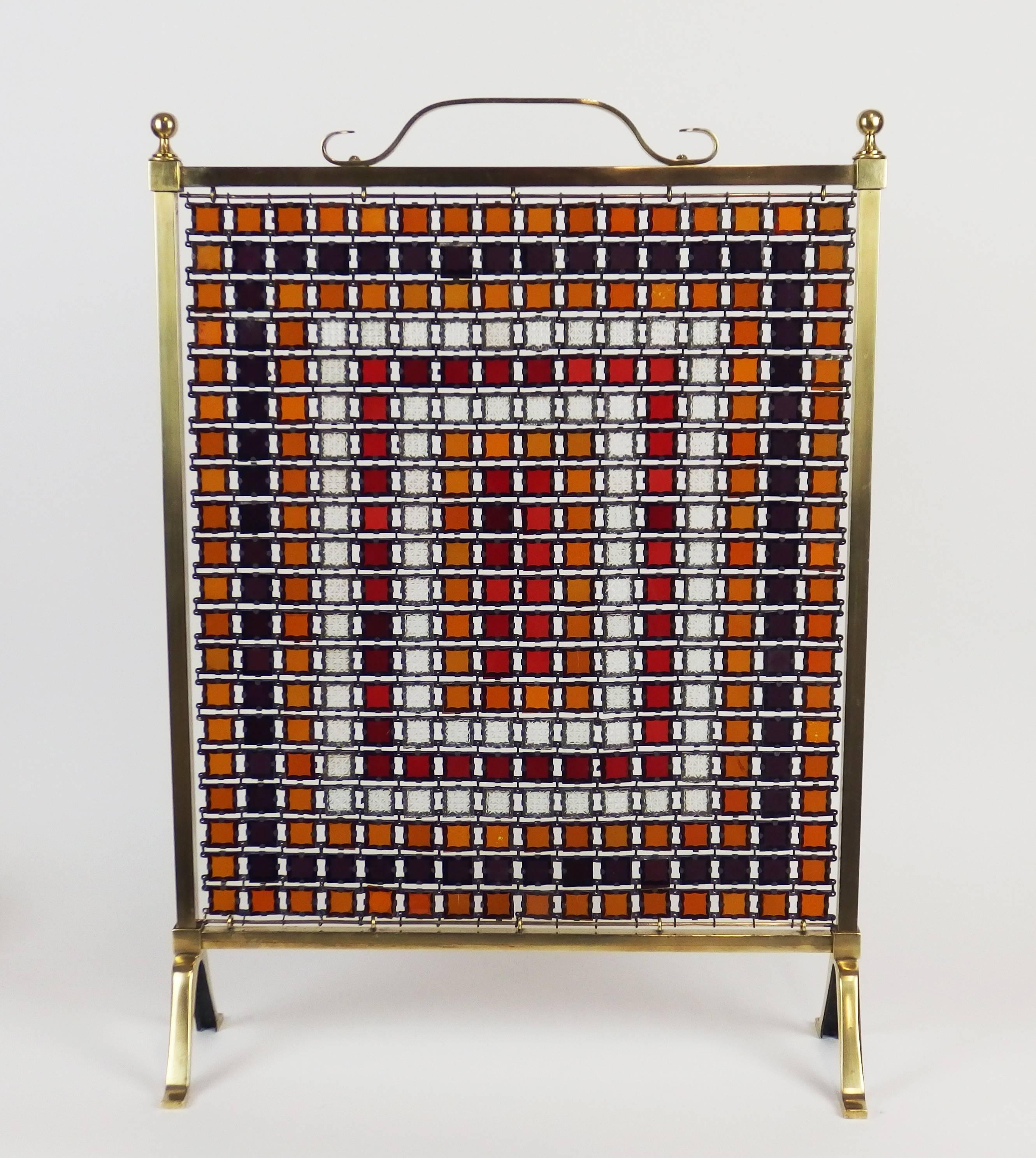 A chain mail fire screen with red, yellow, purple, white glass tiles inserted in zinc frames, hung in a brass and bronze frame. Unidentified stamp under the base.