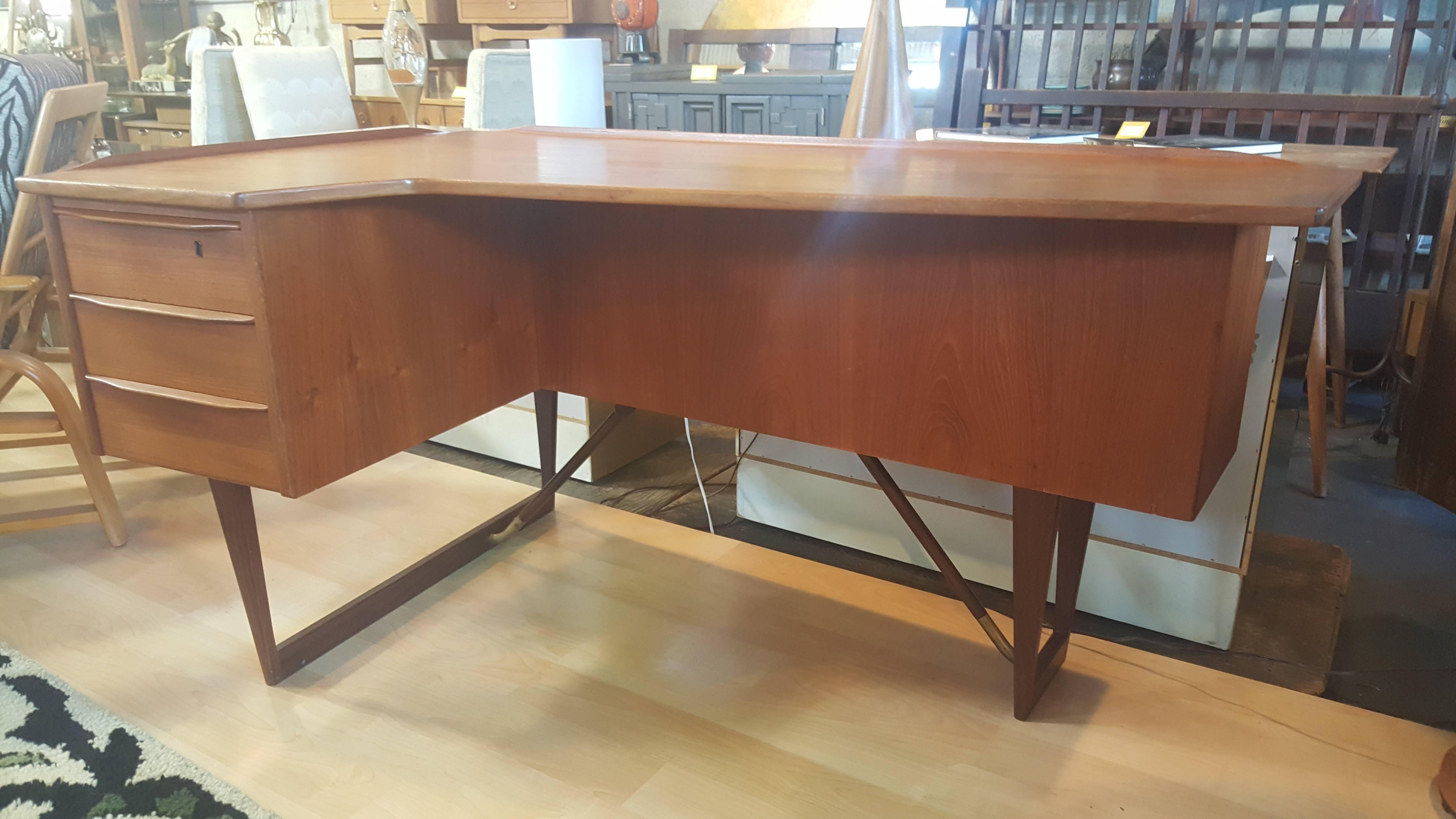 A teak Danish Modern desk designed by Peter Lovig Neilsen, Denmark, 1950s. Boomerang-shaped desk top with molded edges. Reverse side features a locking mirrored bar and bookshelf. Notice turned teak leg supports with brass fittings. Key included.