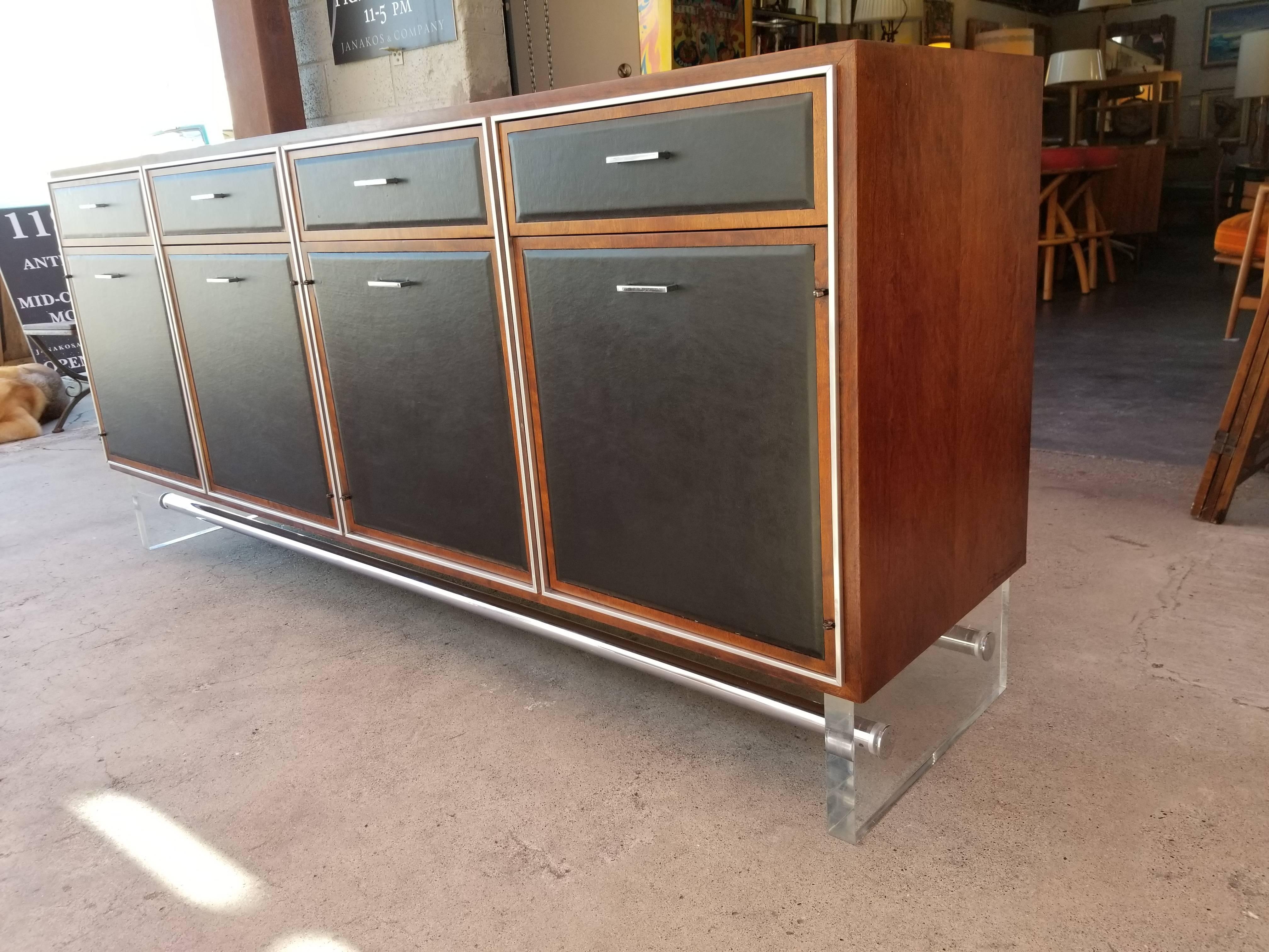 A unique and substantial 1970s Mid-Century Modern credenza crafted in a figured walnut with black leather detail floating on an acrylic and chrome base. Four drawers and four cabinet doors offer ample storage. Solid oak secondary woods. Nice