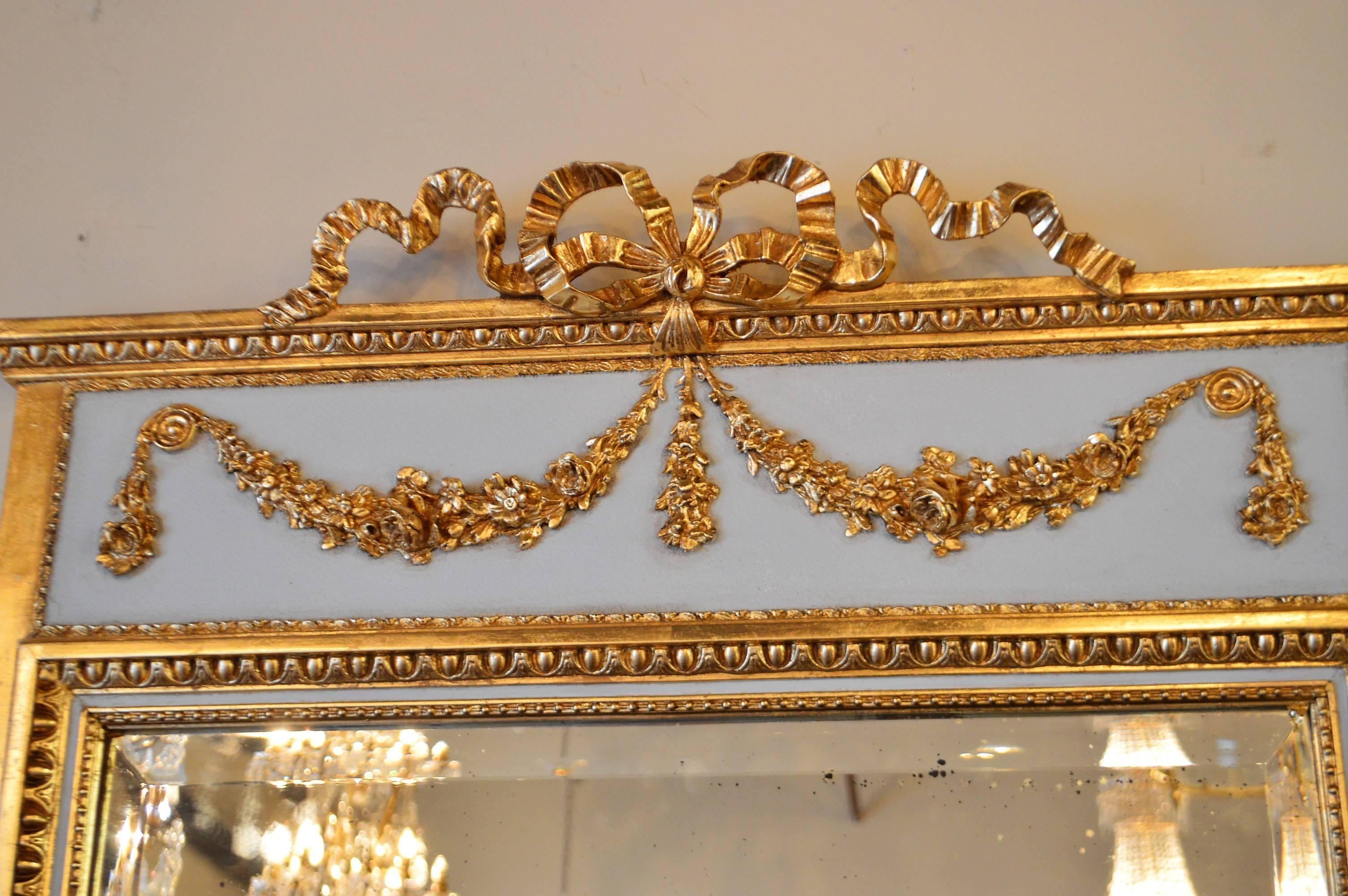 Elegant Louis XVI style trumeau mirror painted and gilt over wood, attractive pale blue is stunning next to the bright gilding. The decorative details including the Louis XVI style bow are all hand-carved. The mirror shows some age.