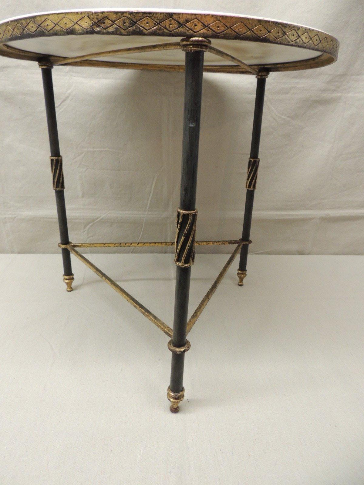 Round Hollywood Regency metal side table, black finish metal with guilt details, mirror top. Three leg table with cross bars legs forming a triangle. Flat black finish with gold leaf details. Diamond and dot pattern apron on the table leading top