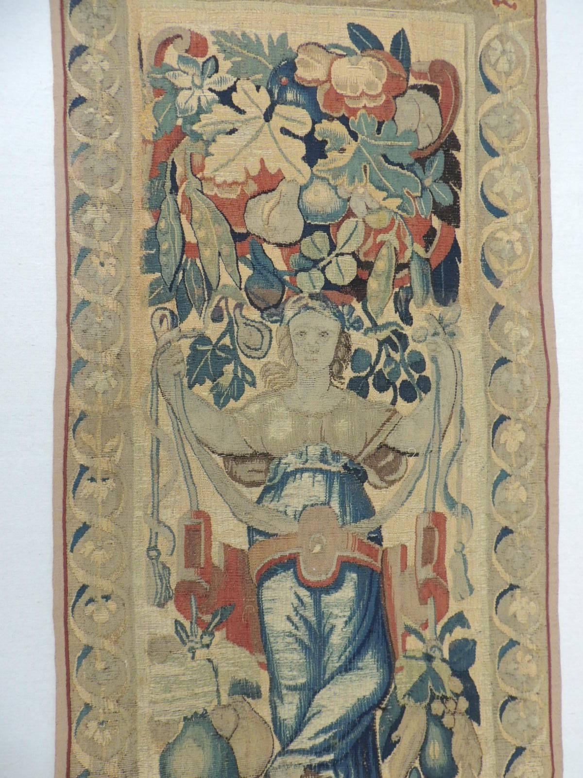 18th century Aubusson tapestry wall-hanging panel.
Textile depicts the autumn with center Goddess holding ribbons surrounded by blooming leaves, fruits and vegetables. Scene is framed by a garland border and linen trim; accentuated by a red silk